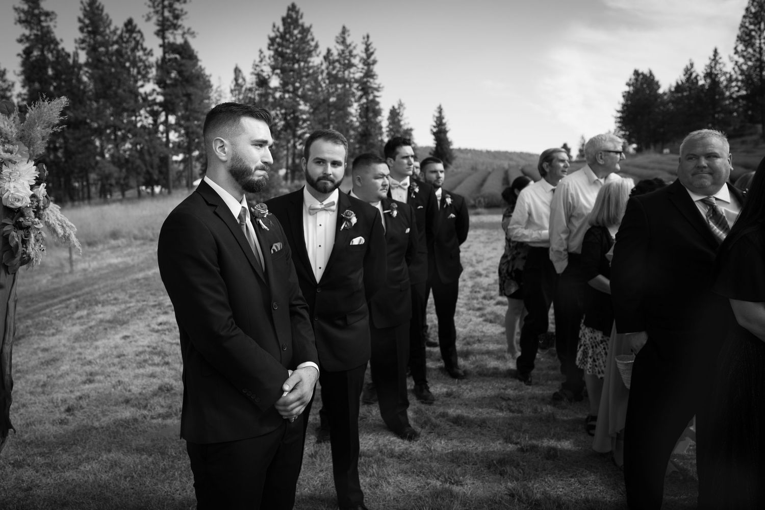 Black and white wedding scene with suited men gathered outdoors among evergreen trees.