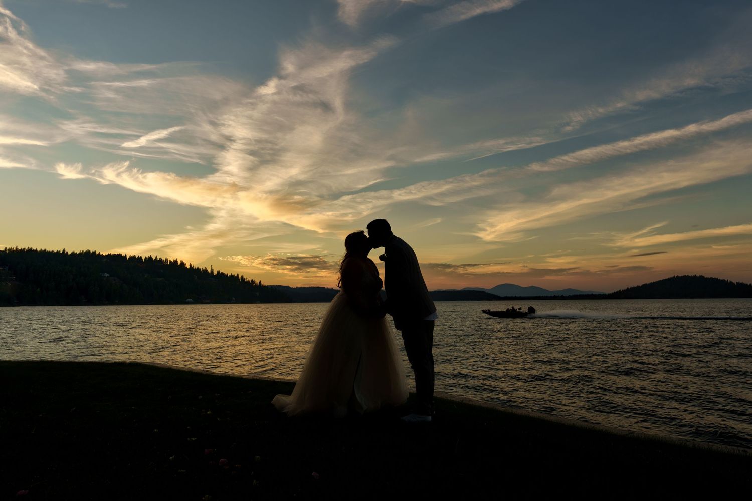 Couple's silhouette against waterfront sunset with dramatic clouds and mountains.