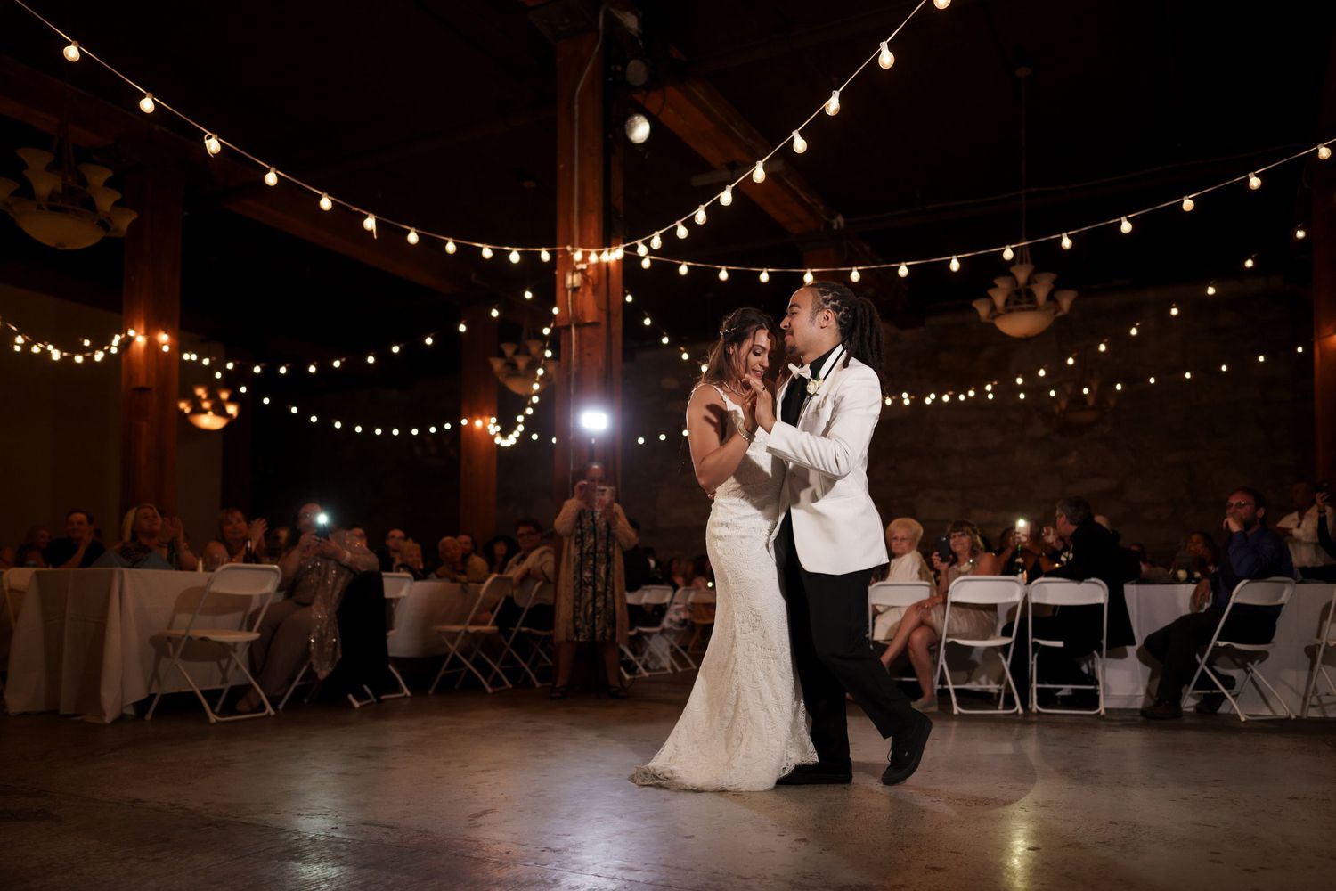Couple sharing first dance under romantic string lights at indoor wedding reception.