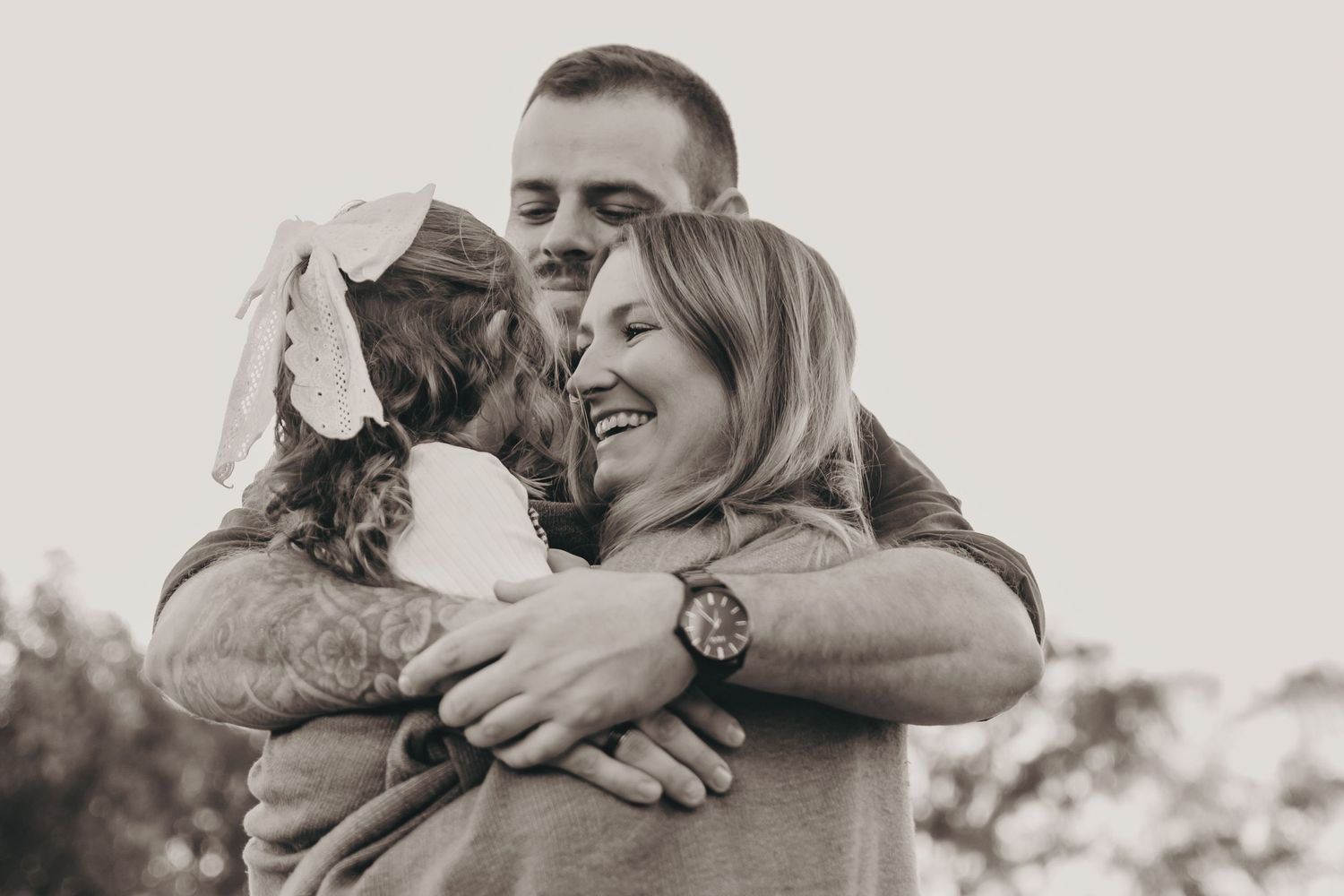 Black and white photo of parents sharing joyful group hug with young daughter outdoors.