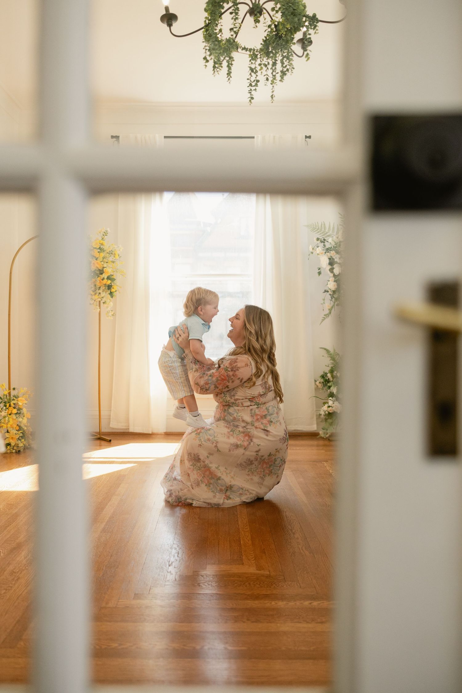 Mother in floral dress kneels on hardwood floor, lovingly holding toddler near sunlit window with greenery decor.
