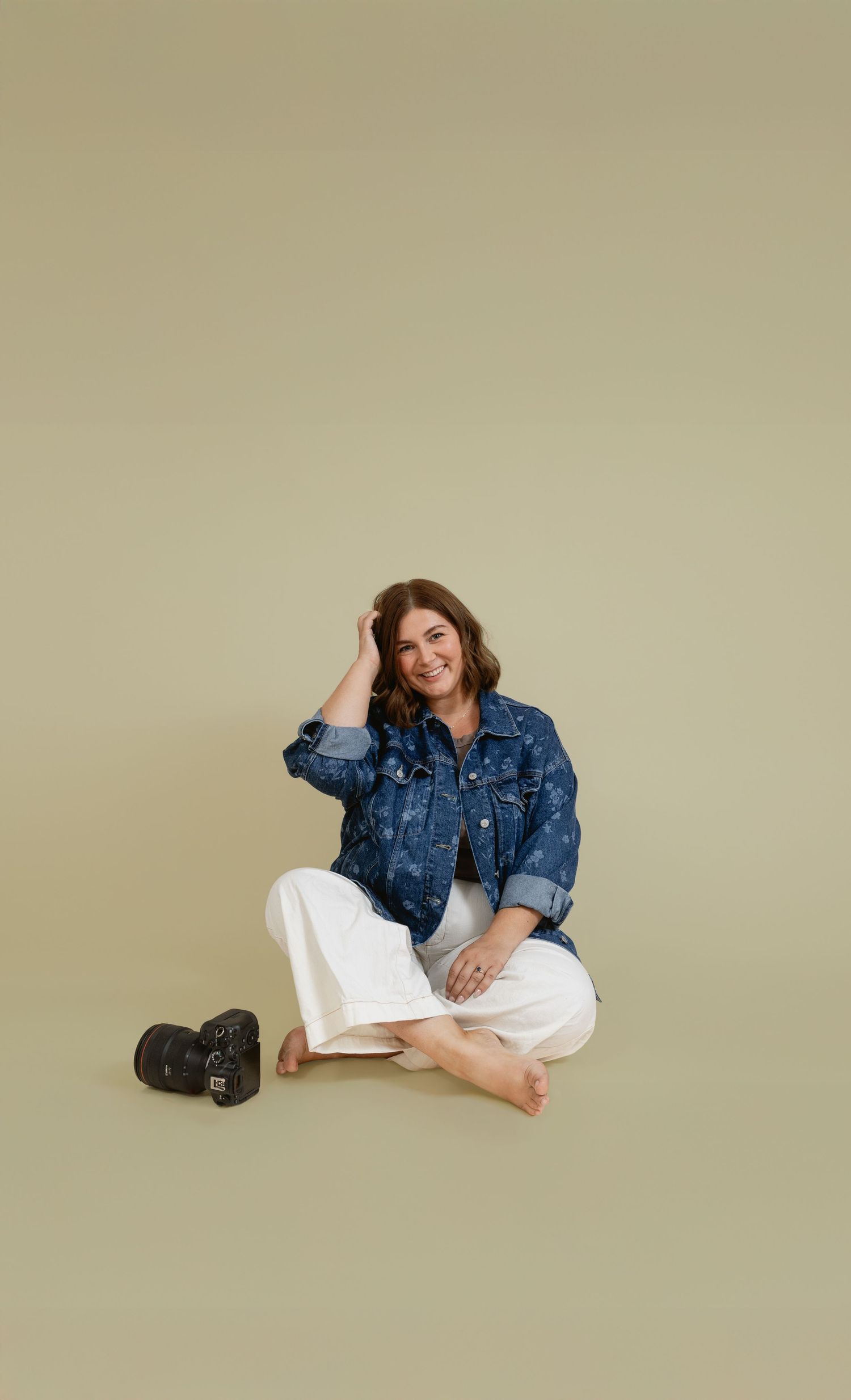 Woman in denim jacket and white pants sits cross-legged on floor smiling, with camera beside her on neutral background.