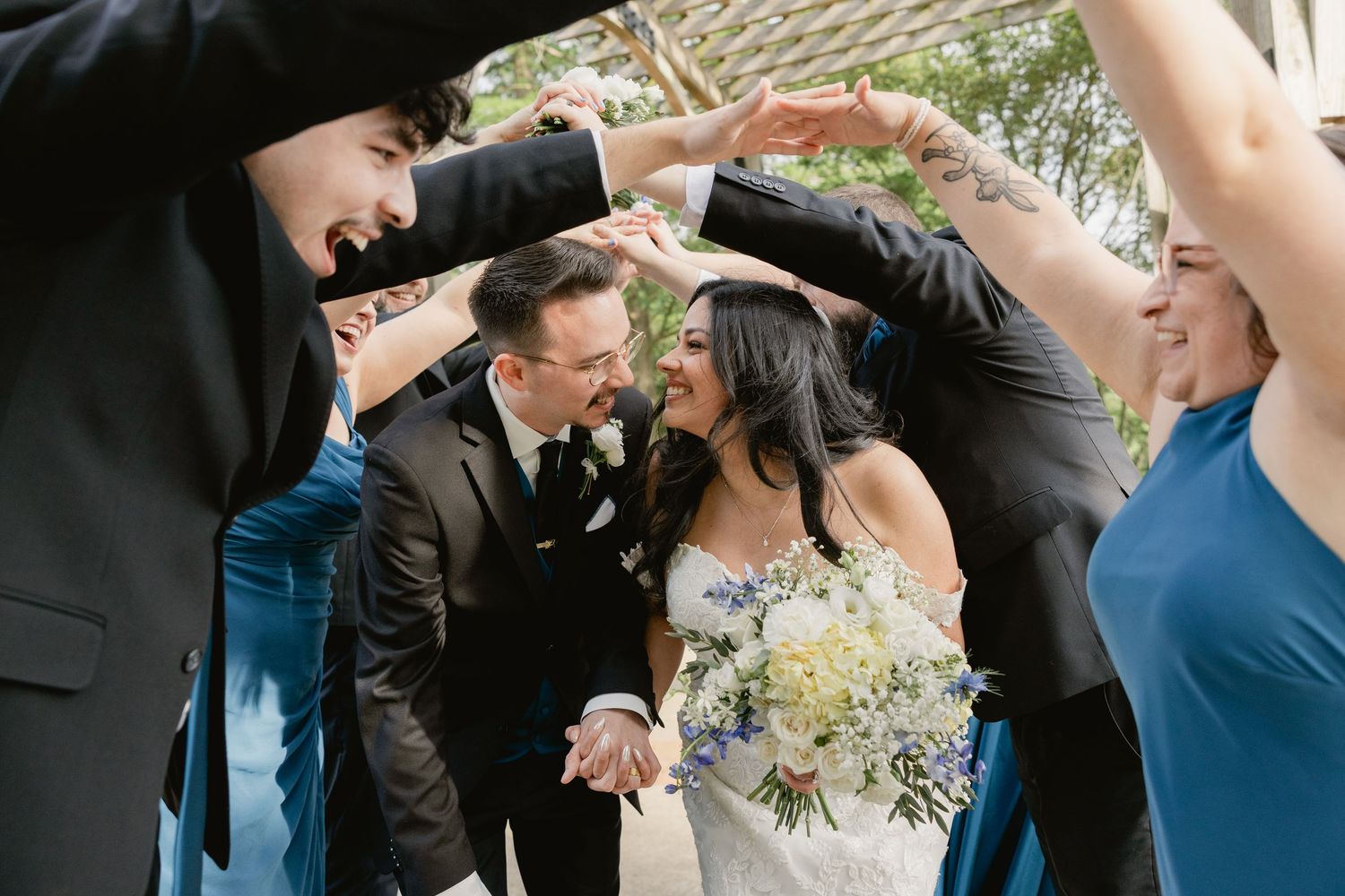 Bride and groom walk through arch of raised hands formed by wedding party in blue attire outdoors.