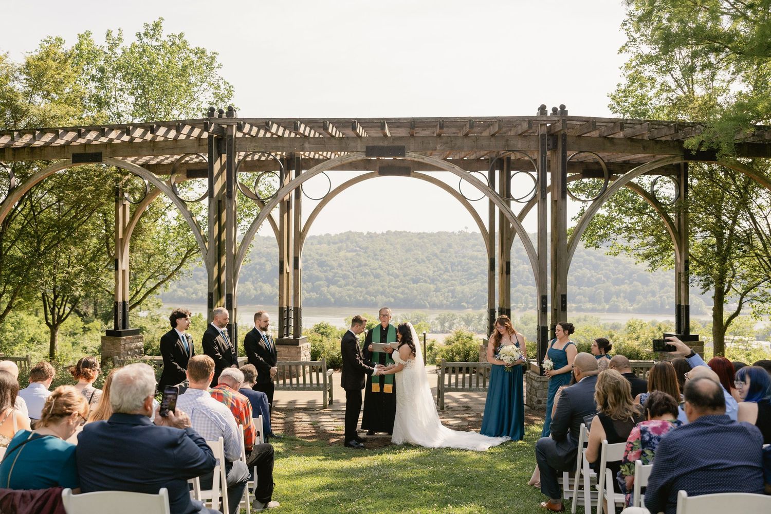 Outdoor wedding ceremony under a wooden pergola overlooking scenic hills, with guests seated on white chairs.