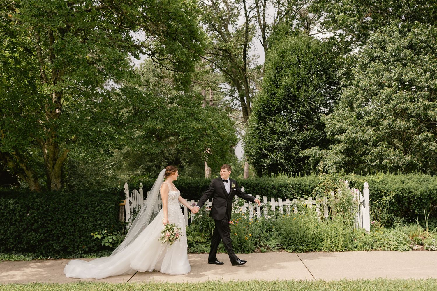 Bride and groom walk hand in hand through a lush garden path with white picket fence on their wedding day.