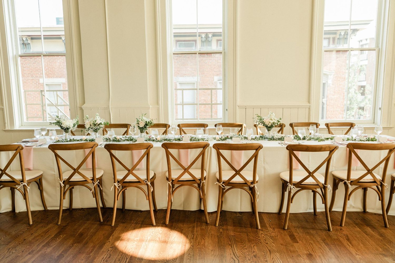 Elegant wedding head table with cross-back wooden chairs, white floral centerpieces, and tall windows in a bright venue.