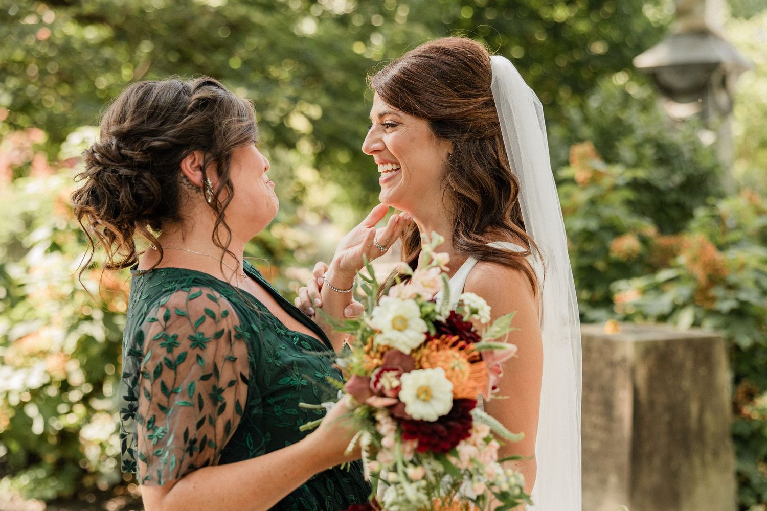 Bride in white gown and veil laughs with bridesmaid in green floral dress while holding bouquet outdoors.