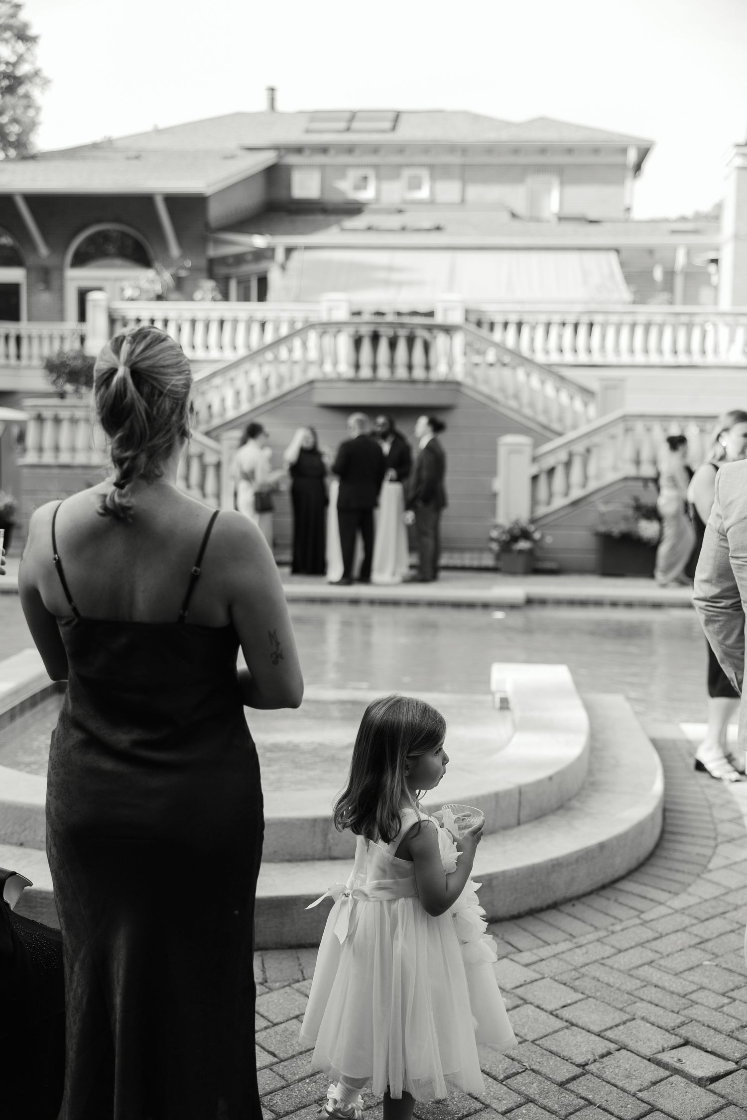 Black and white photo showing a woman and young girl in formal dresses watching a gathering by ornate white stairs.Cincinnati Wedding Photog