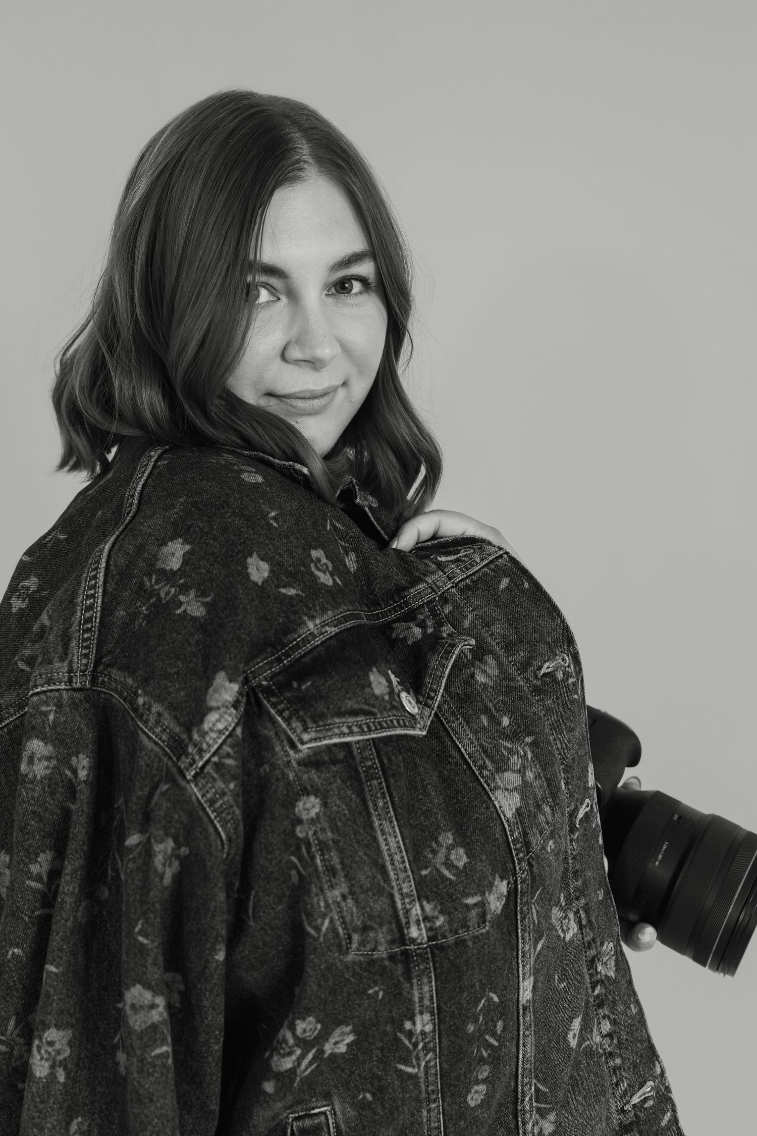 Black and white portrait of a young woman holding a camera, wearing a floral denim jacket, smiling over her shoulder.