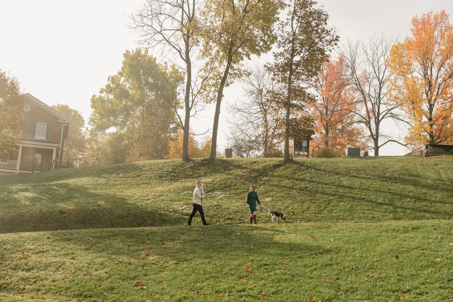 Two people walk a dog on a lush green hillside park surrounded by autumn trees on a misty morning.