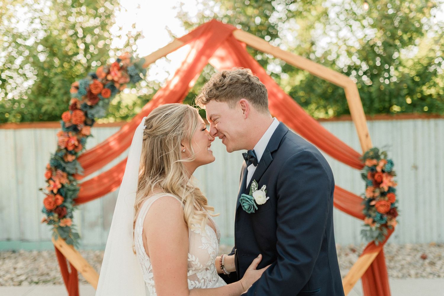 A romantic wedding ceremony takes place under a coral-colored arch decorated with flowers in an outdoor garden setting.