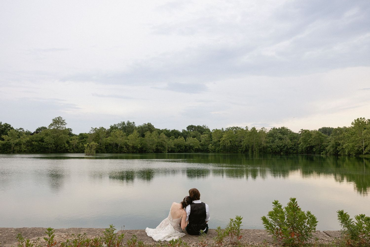 Bride and groom sitting together by a serene lake surrounded by lush green trees on their wedding day.