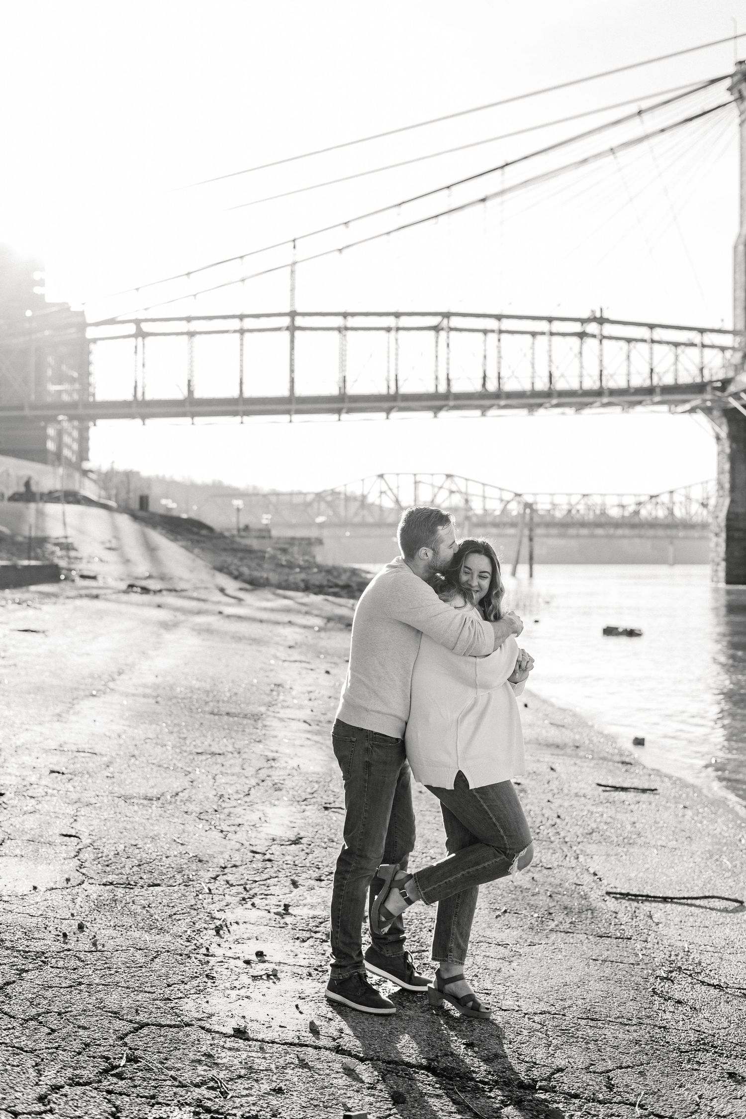Couple embracing near riverside bridge on misty morning in black and white.