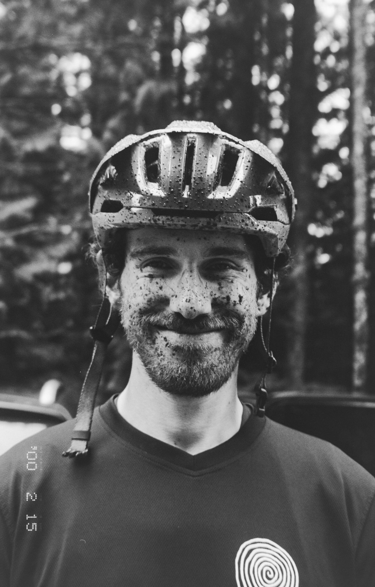 A mountain biker wearing a helmet and covered in dirt smiles at the camera in a forest setting.