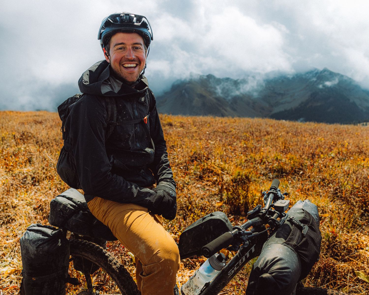 Mountain biker takes a break in autumn meadow with misty peaks in background, wearing black jacket and yellow pants.