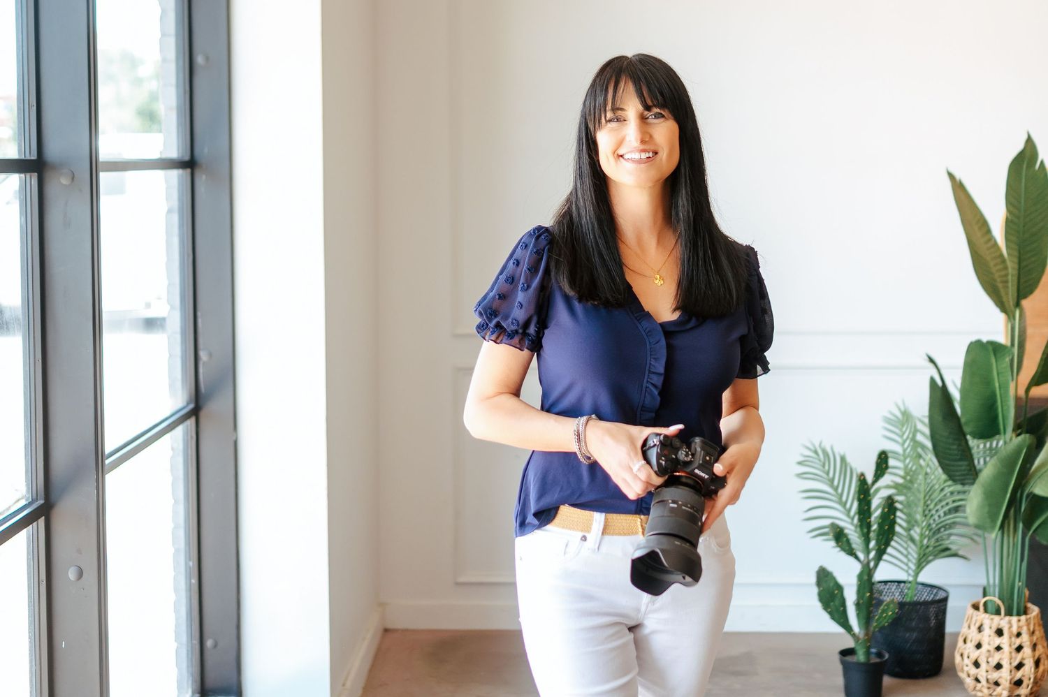 Smiling woman with dark hair holding a camera in a bright studio with white walls and green plants.
