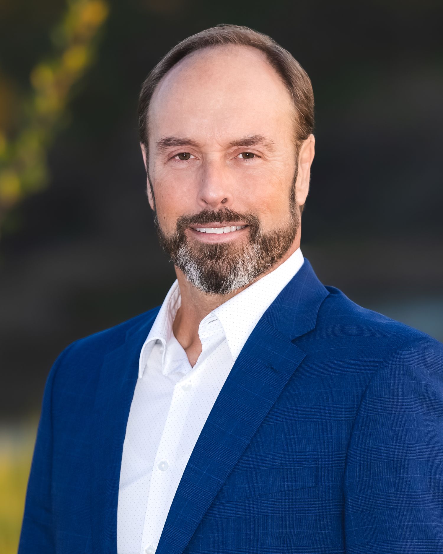 Professional headshot of a smiling man with a beard wearing a blue blazer and white shirt outdoors.