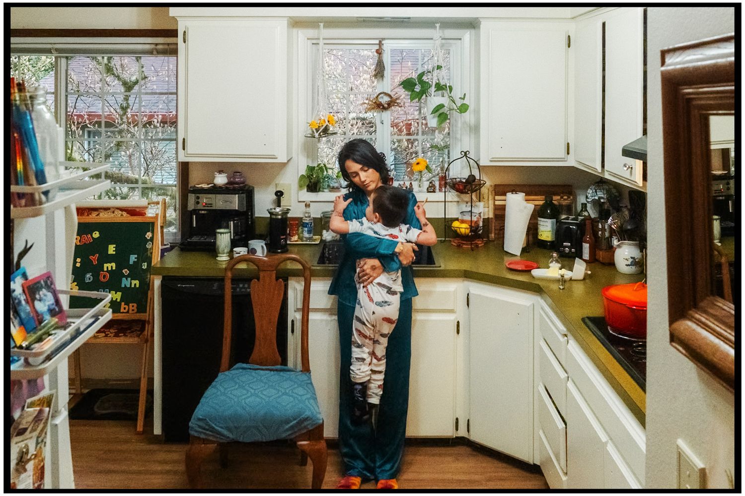 A person wearing a navy jumpsuit stands in a kitchen with white cabinets and colorful artwork on the walls.