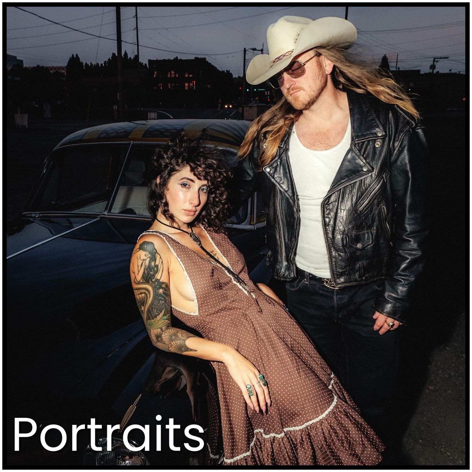 Two people in western-inspired outfits pose at dusk near a vintage car in an artistic portrait shoot.