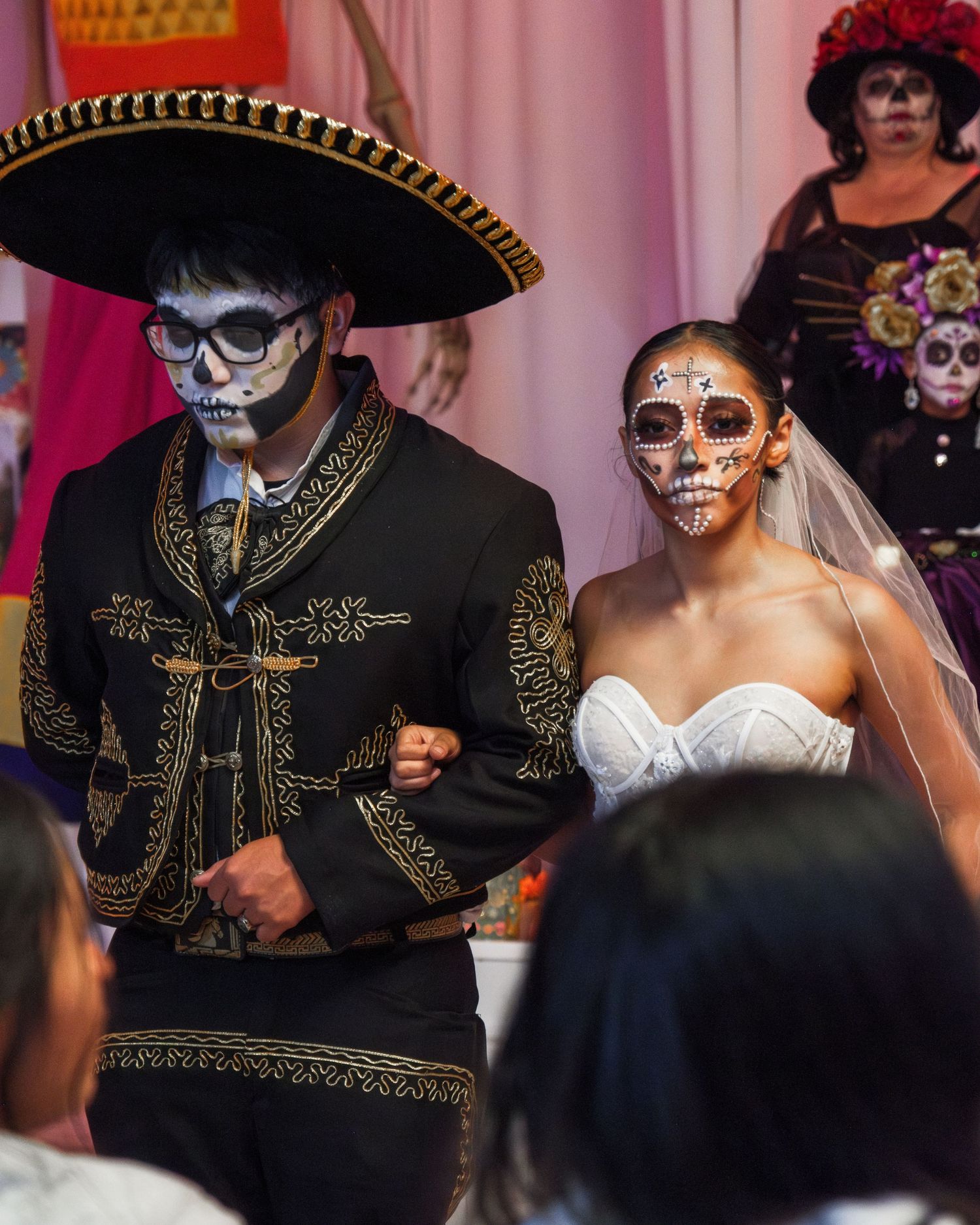 Two people in Dia de los Muertos skull makeup and costumes at a wedding, with bride in white dress and groom in mariachi attire.