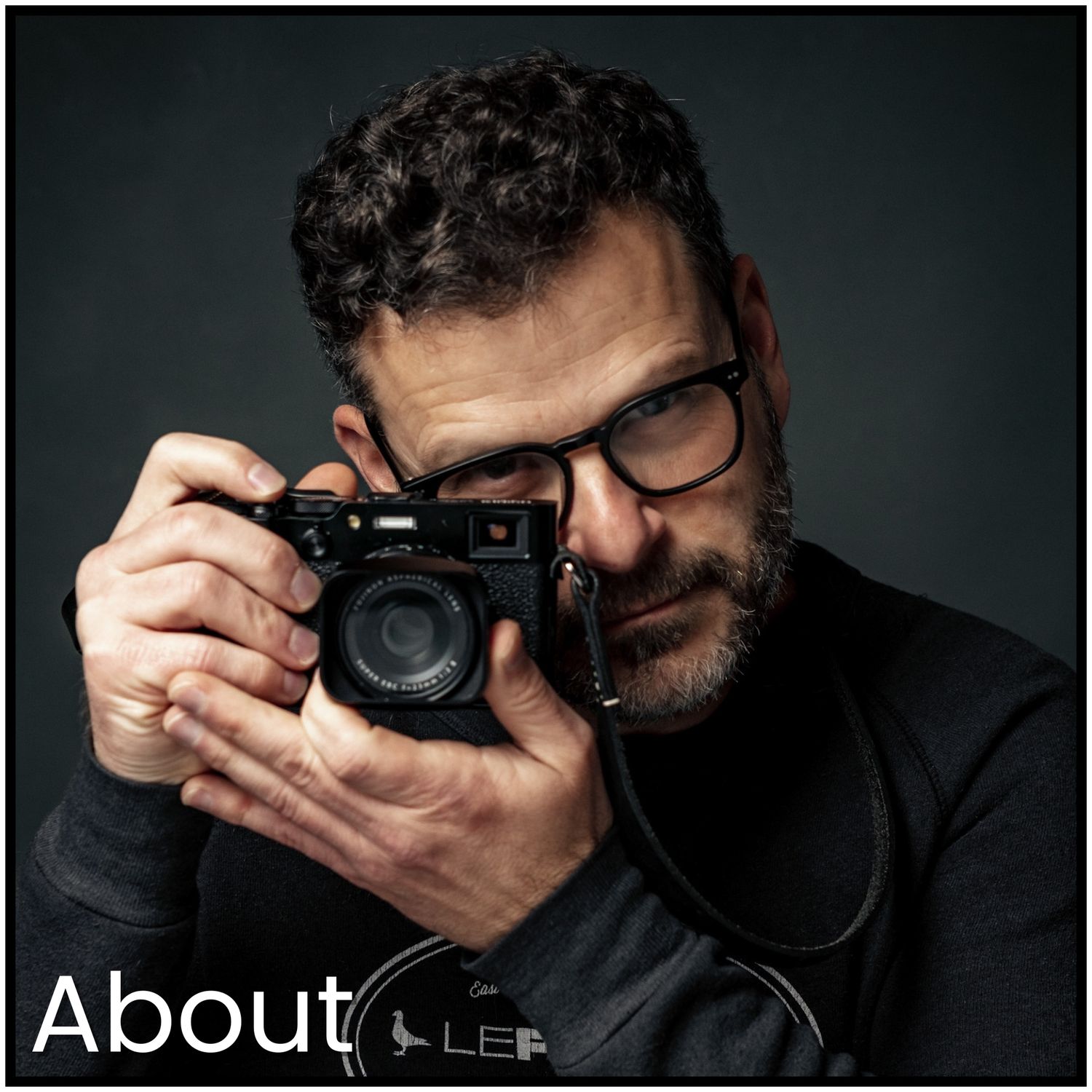 Photographer wearing glasses and black sweater holds vintage film camera against dark background.