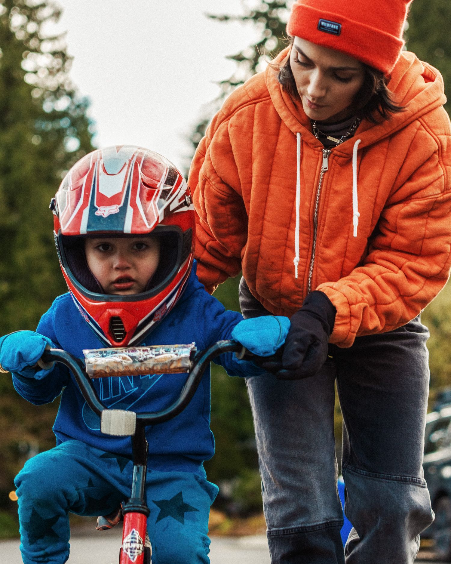 Someone in bright orange winter wear helps a young child learning to ride a bike while wearing a red and blue safety helmet.