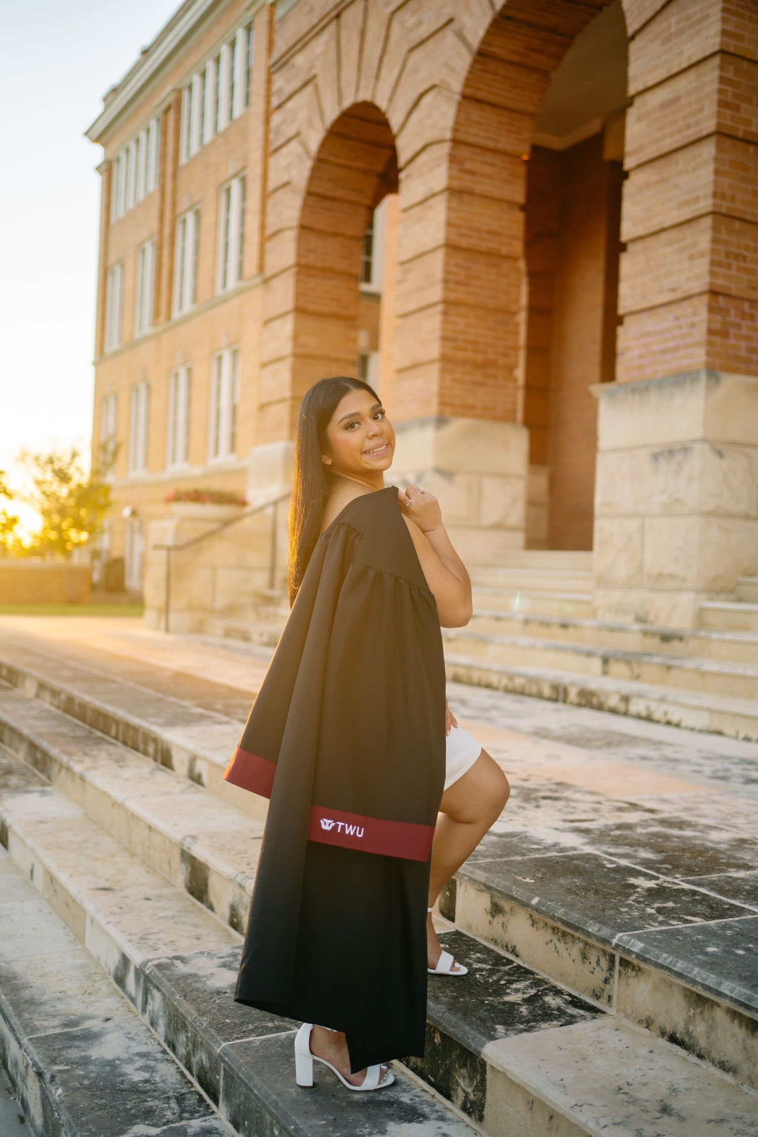 Young woman in TWU graduation gown smiling on stone steps outside a brick university building at golden hour.