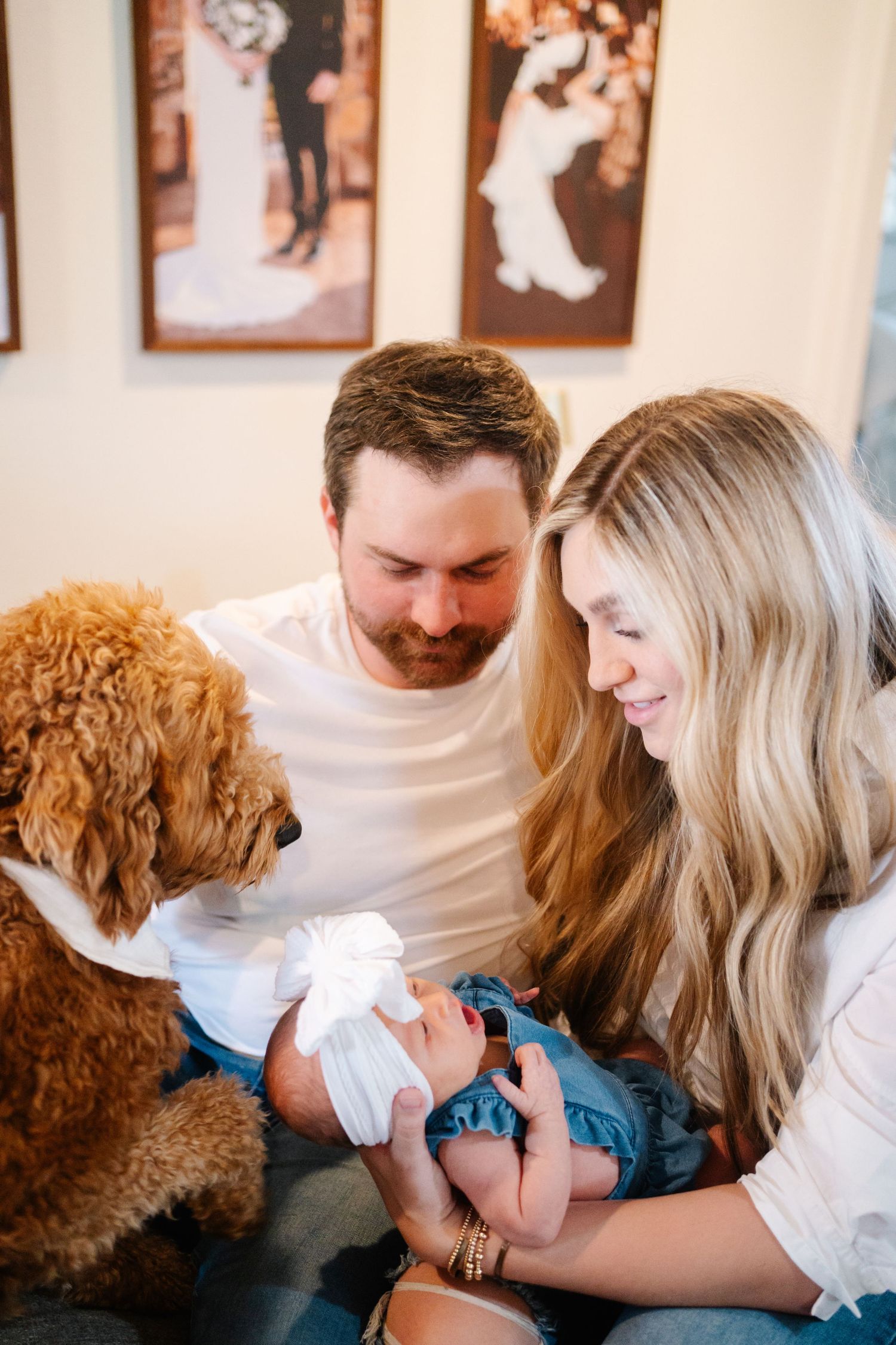 A couple smiles at their newborn baby while a fluffy golden doodle looks on, with wedding photos displayed on the wall.
