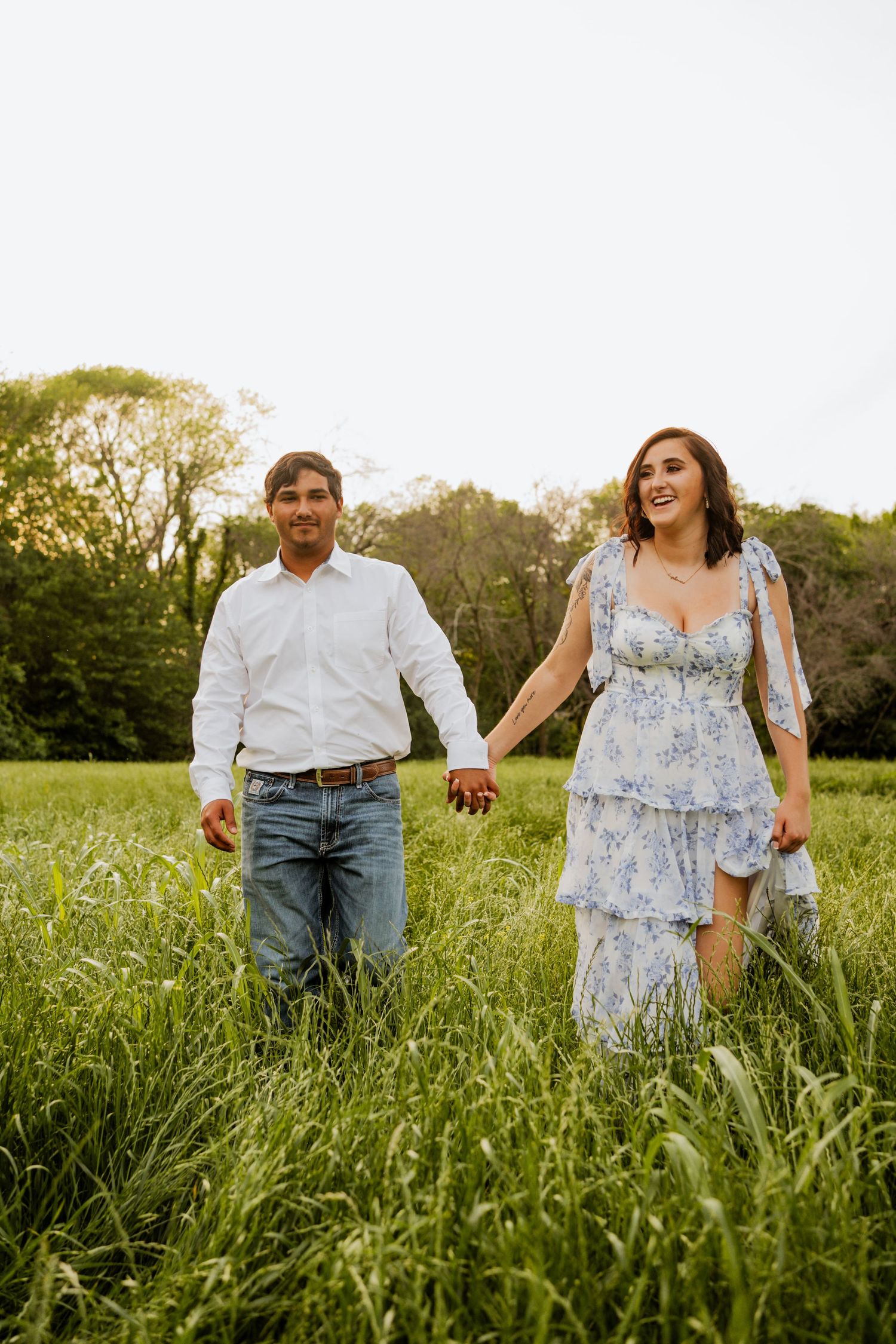 A couple holds hands walking through a lush green field, woman in floral dress, man in white shirt and jeans.