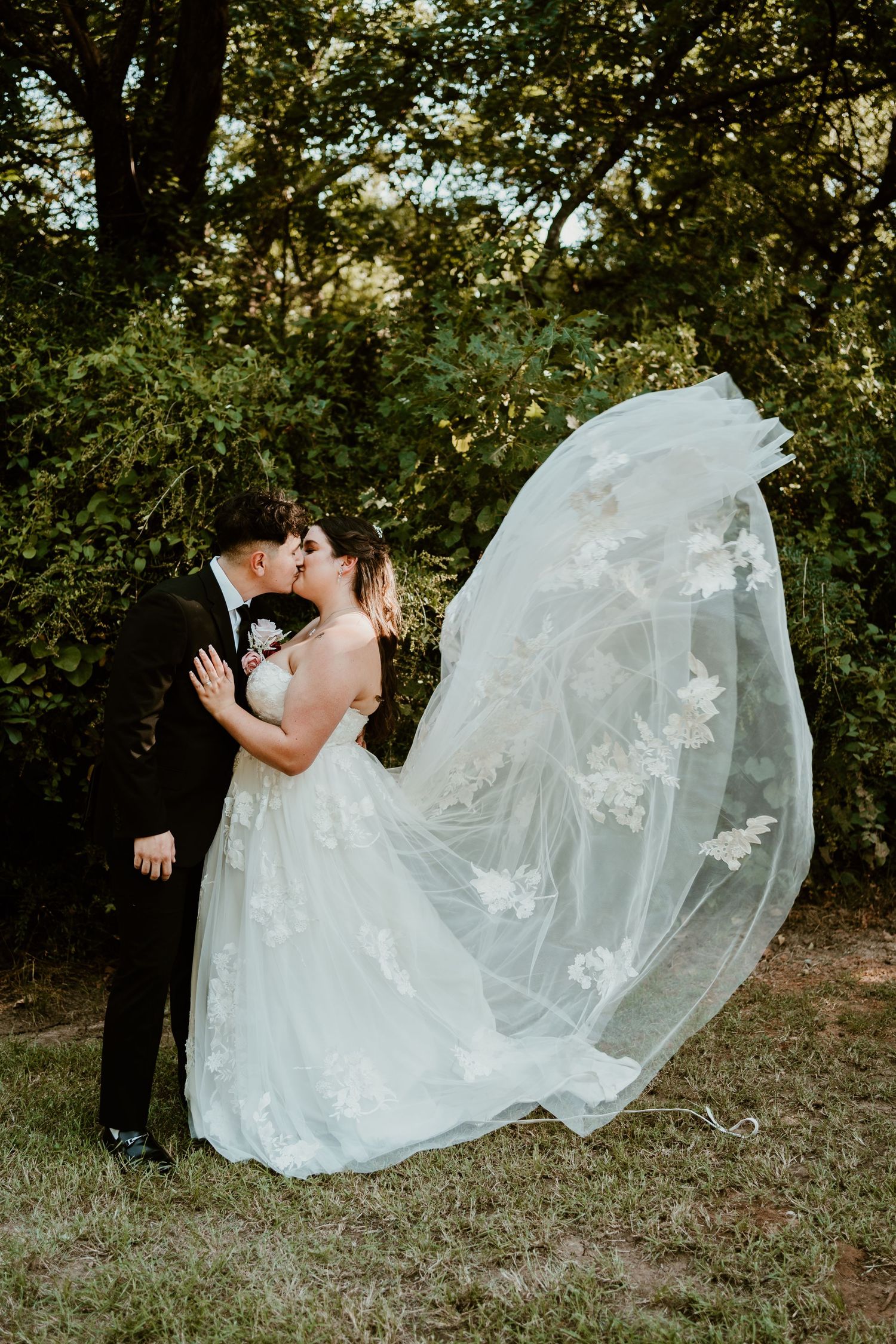 Bride and groom share a kiss outdoors as her lace veil flows dramatically in the wind against green trees.