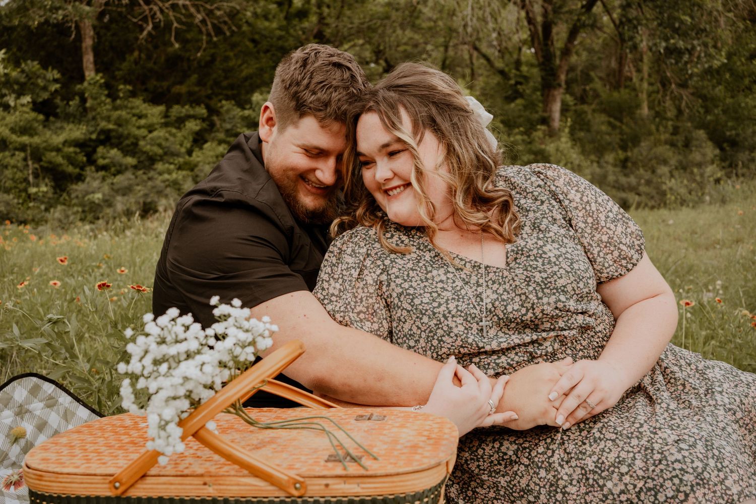 Couple sharing a romantic picnic moment in a wildflower meadow, sitting on a blanket with a wicker basket and white flowers.