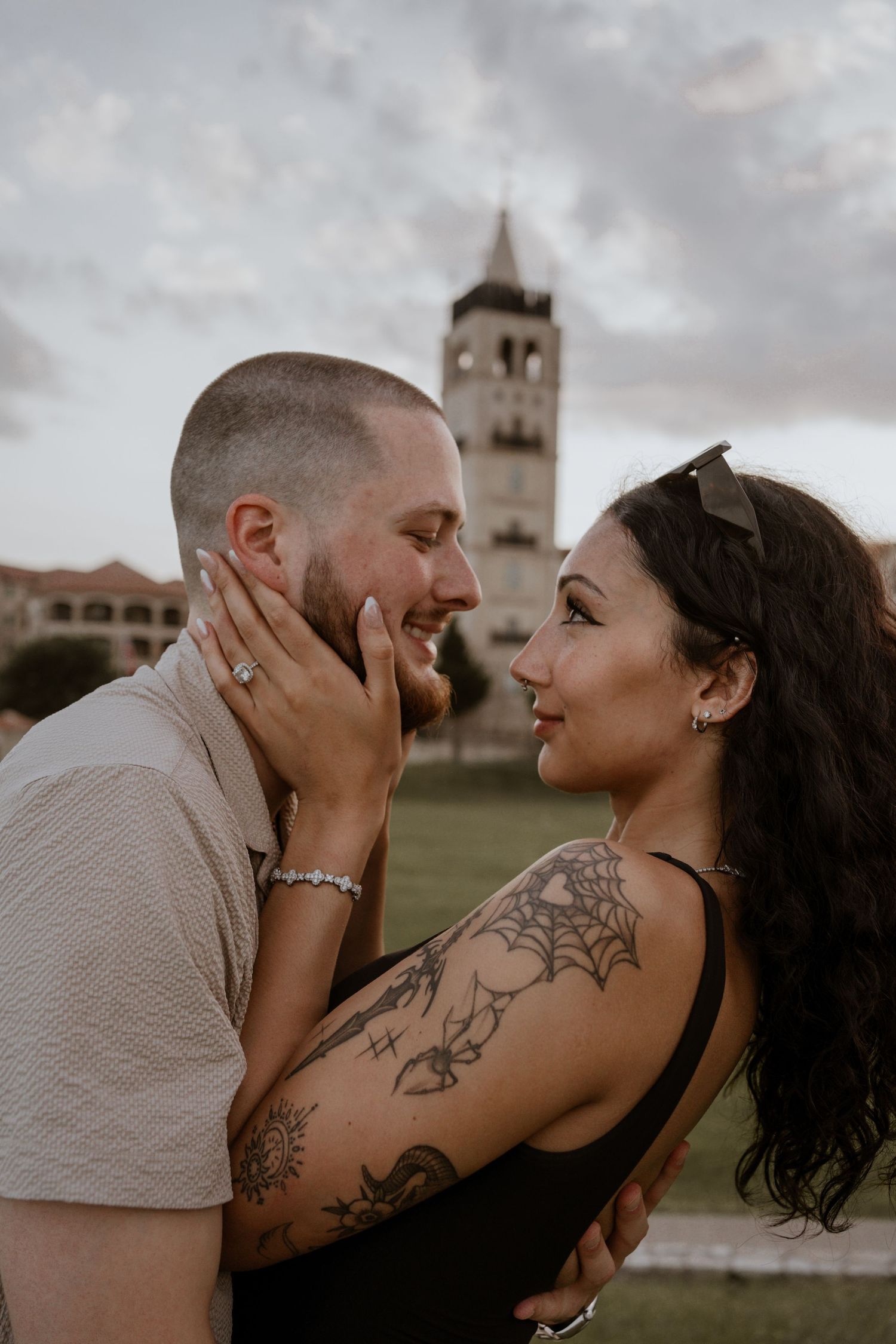 A tattooed couple shares a romantic moment outdoors, with a historic tower building visible in the background.