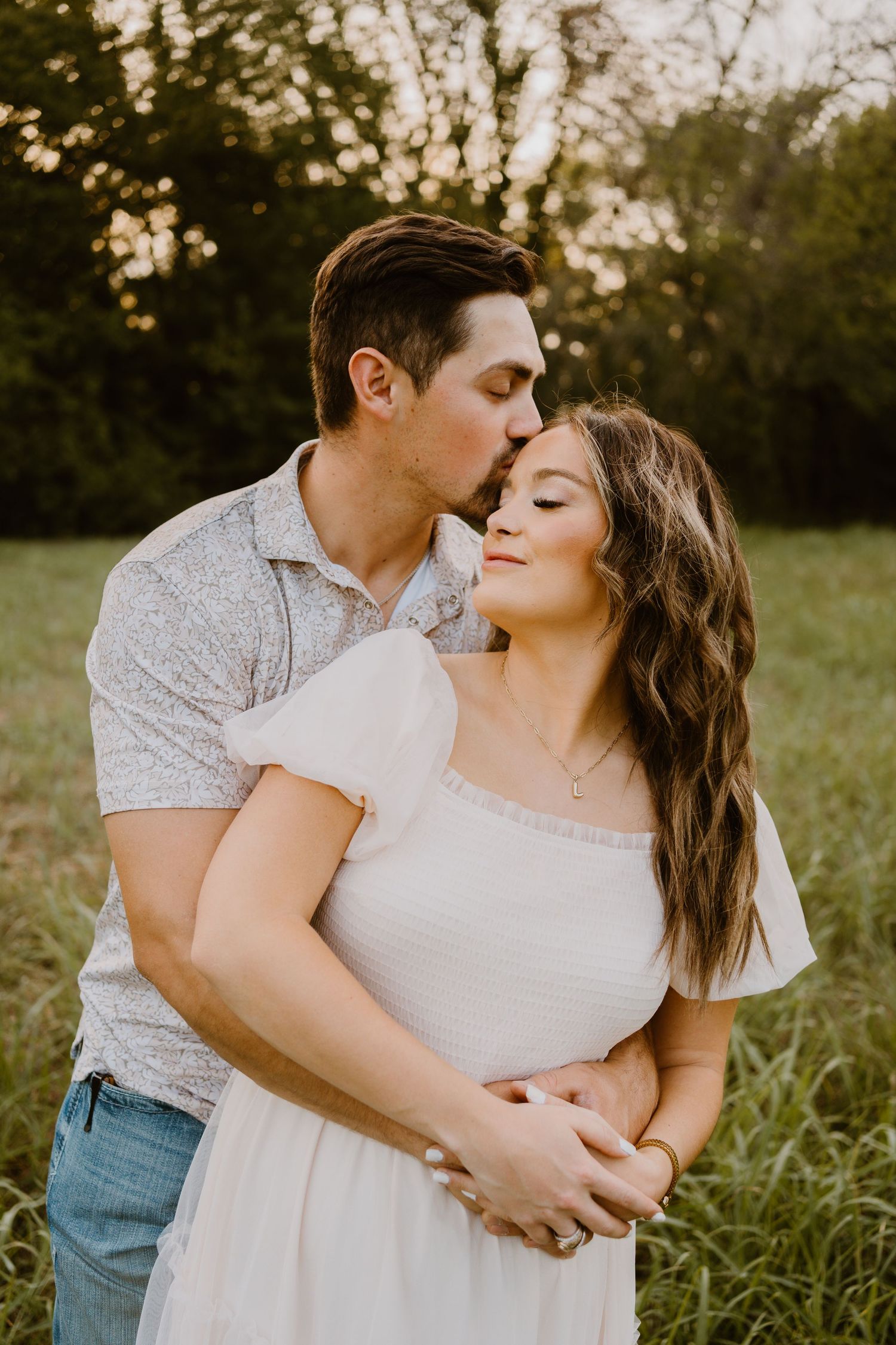 A couple shares a tender moment outdoors, man kissing woman's temple while embracing in a sunlit green field.