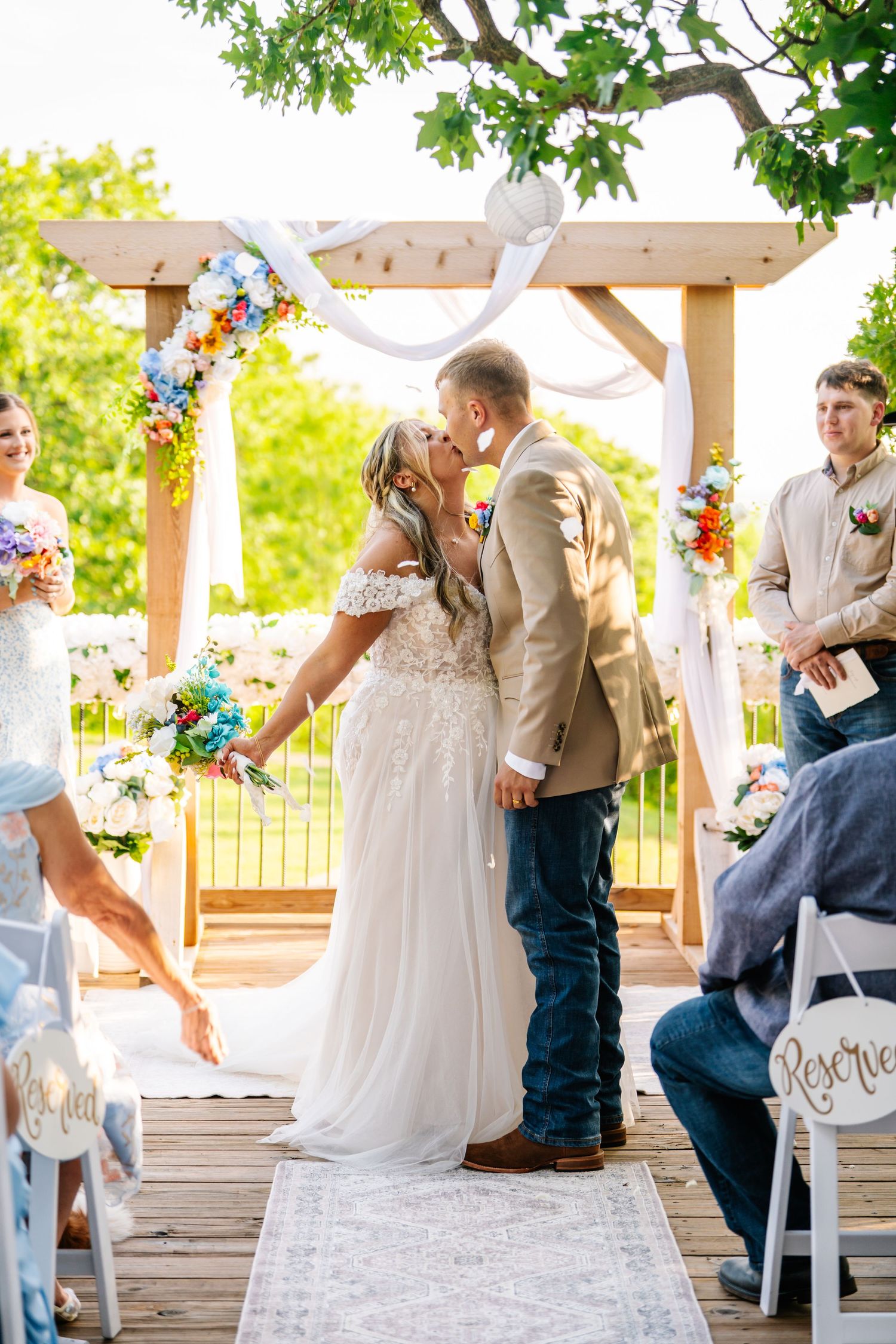 Bride and groom share first kiss under floral wooden arch at outdoor wedding ceremony on sunny day.