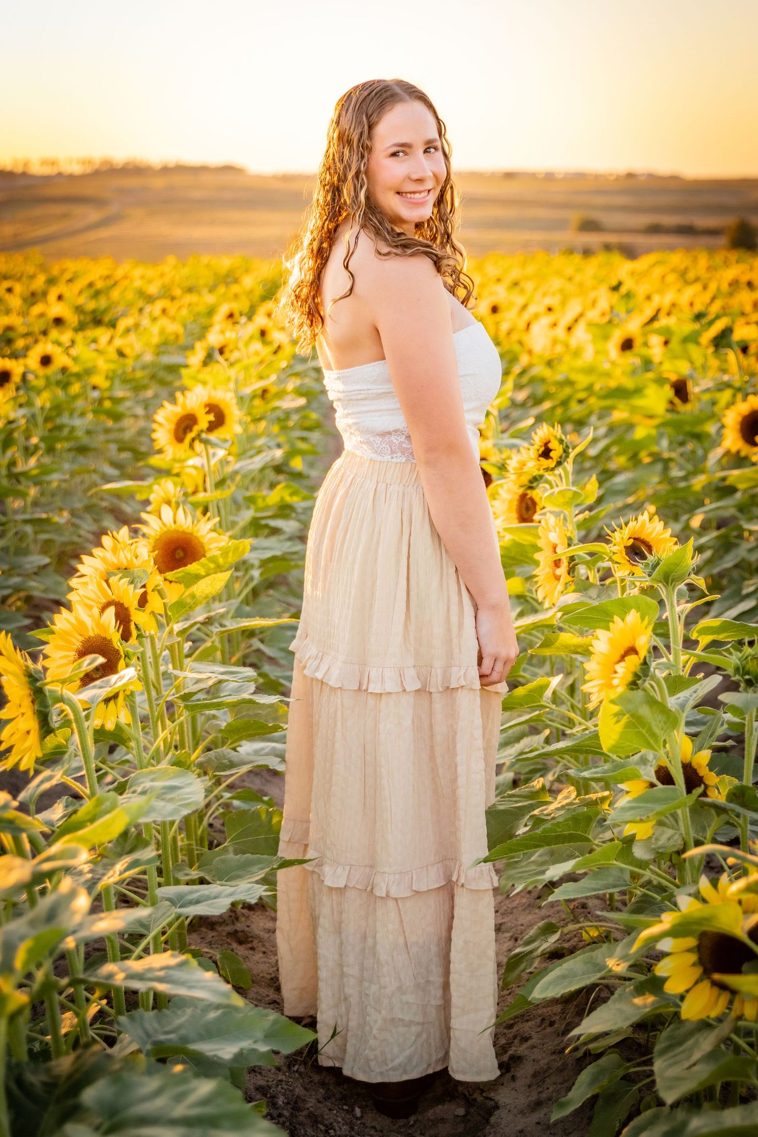 A dreamy golden hour photoshoot in a vibrant sunflower field with a white lace dress creating a romantic summer scene.