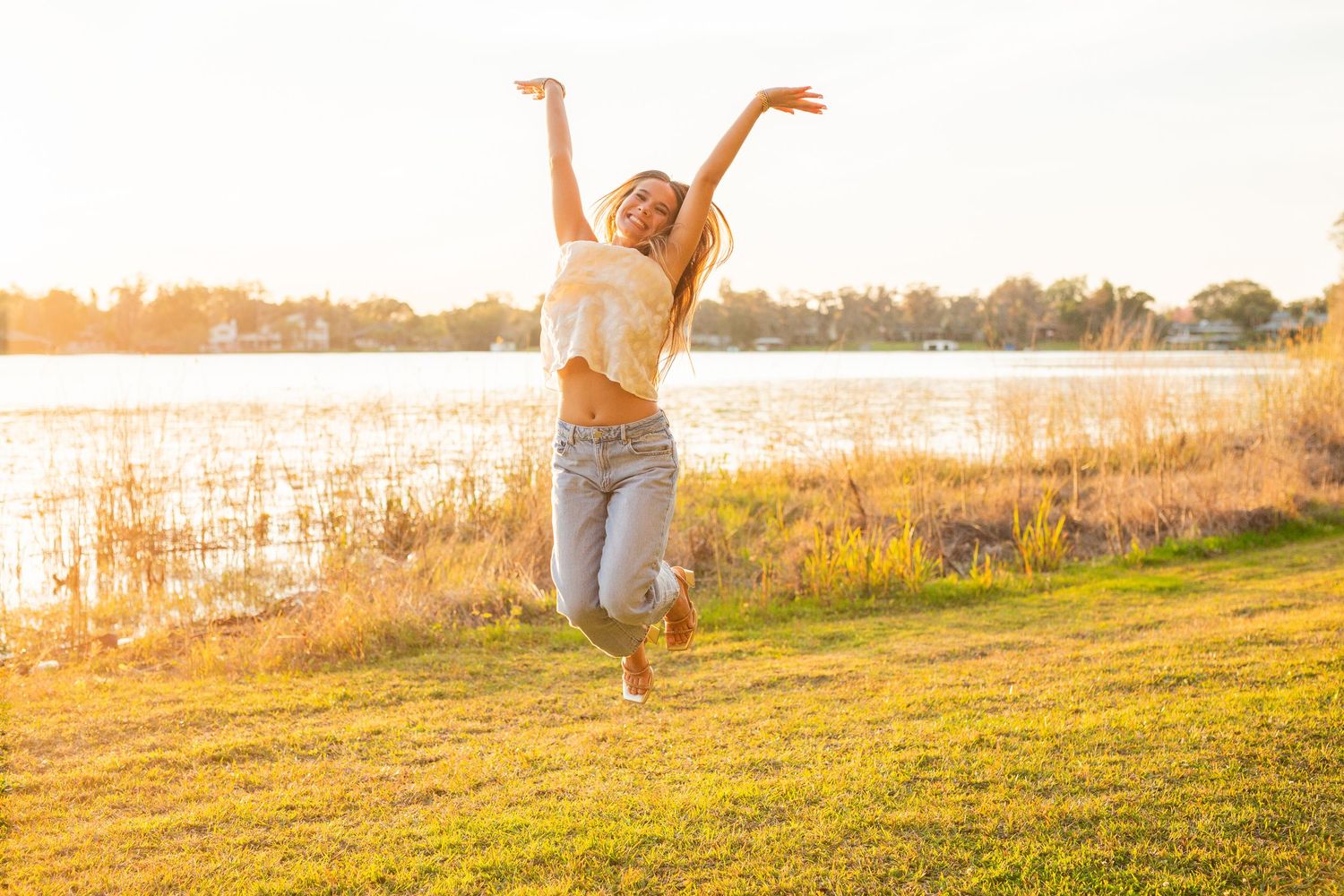 A person joyfully jumps in the air on a grassy hill during golden hour sunset by the water.