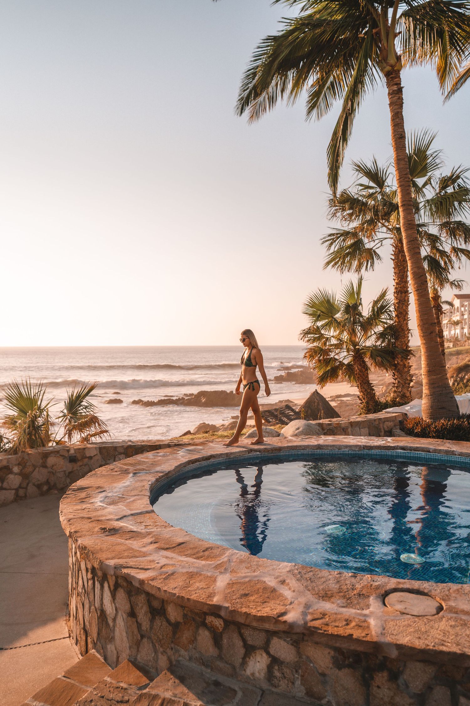 Oceanfront infinity pool surrounded by palm trees at sunset.