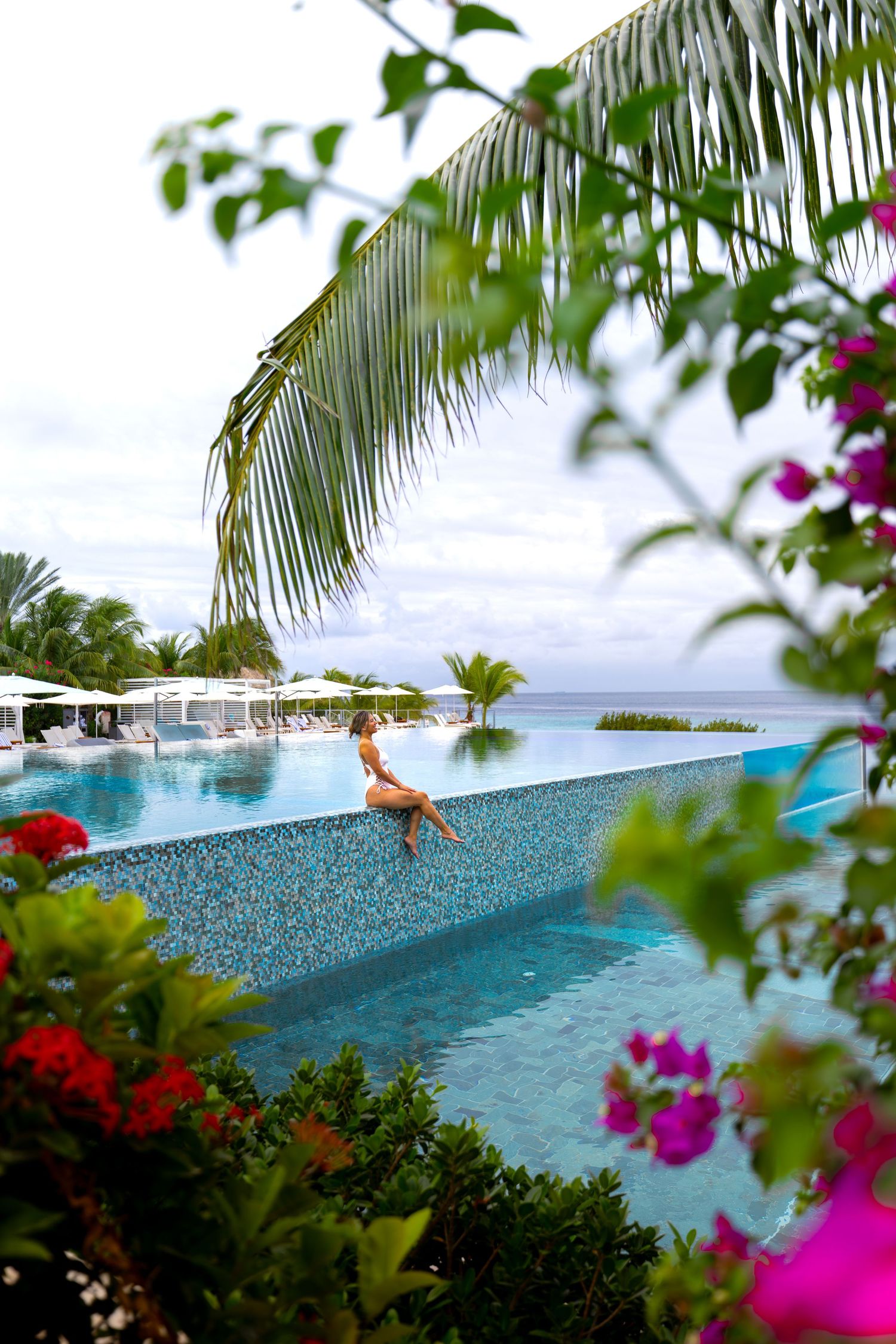 Infinity pool overlooking the ocean with tropical flowers and palm trees framing the view.