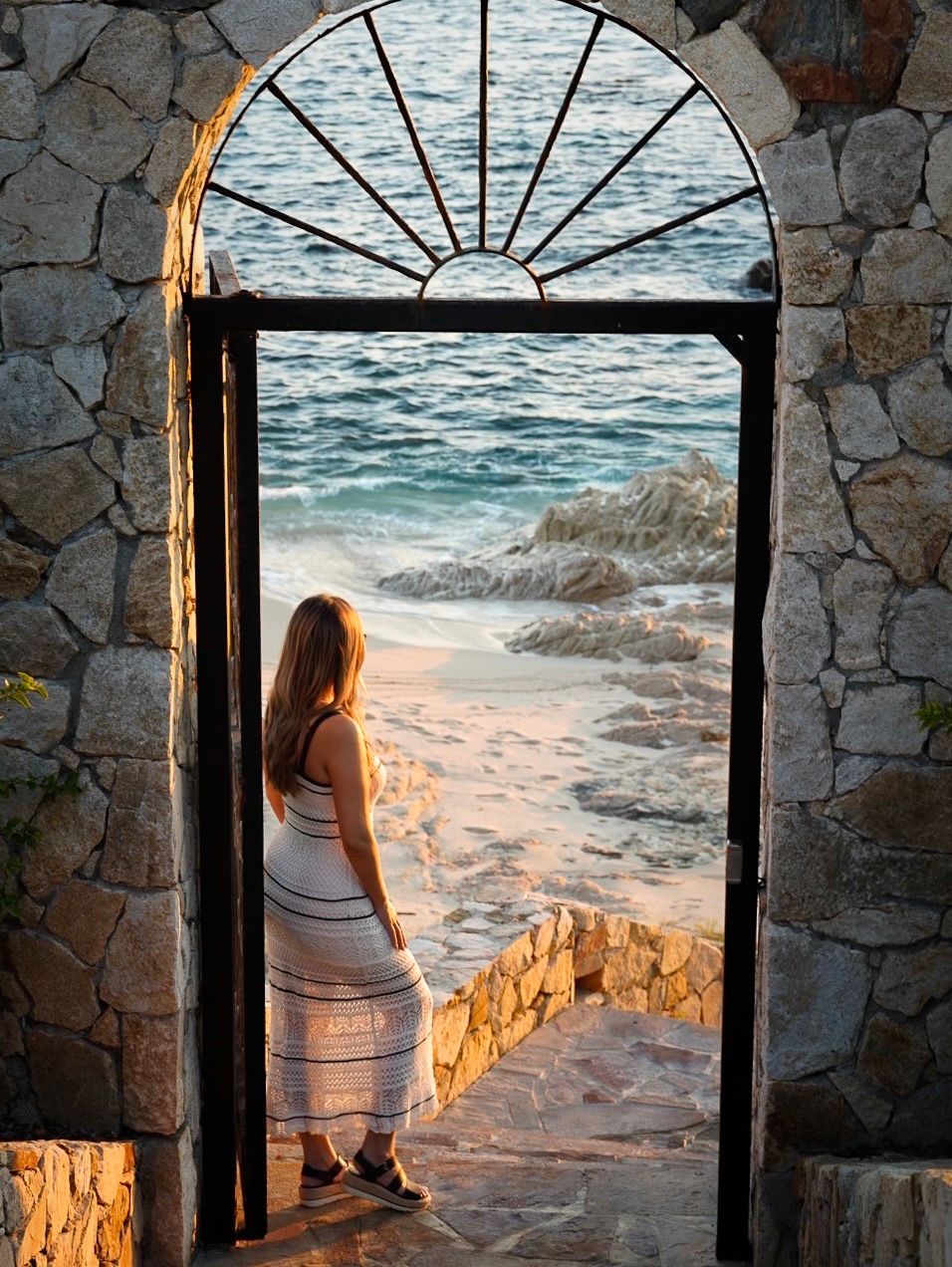 A stone archway frames a view of a beach and ocean waves at sunset.