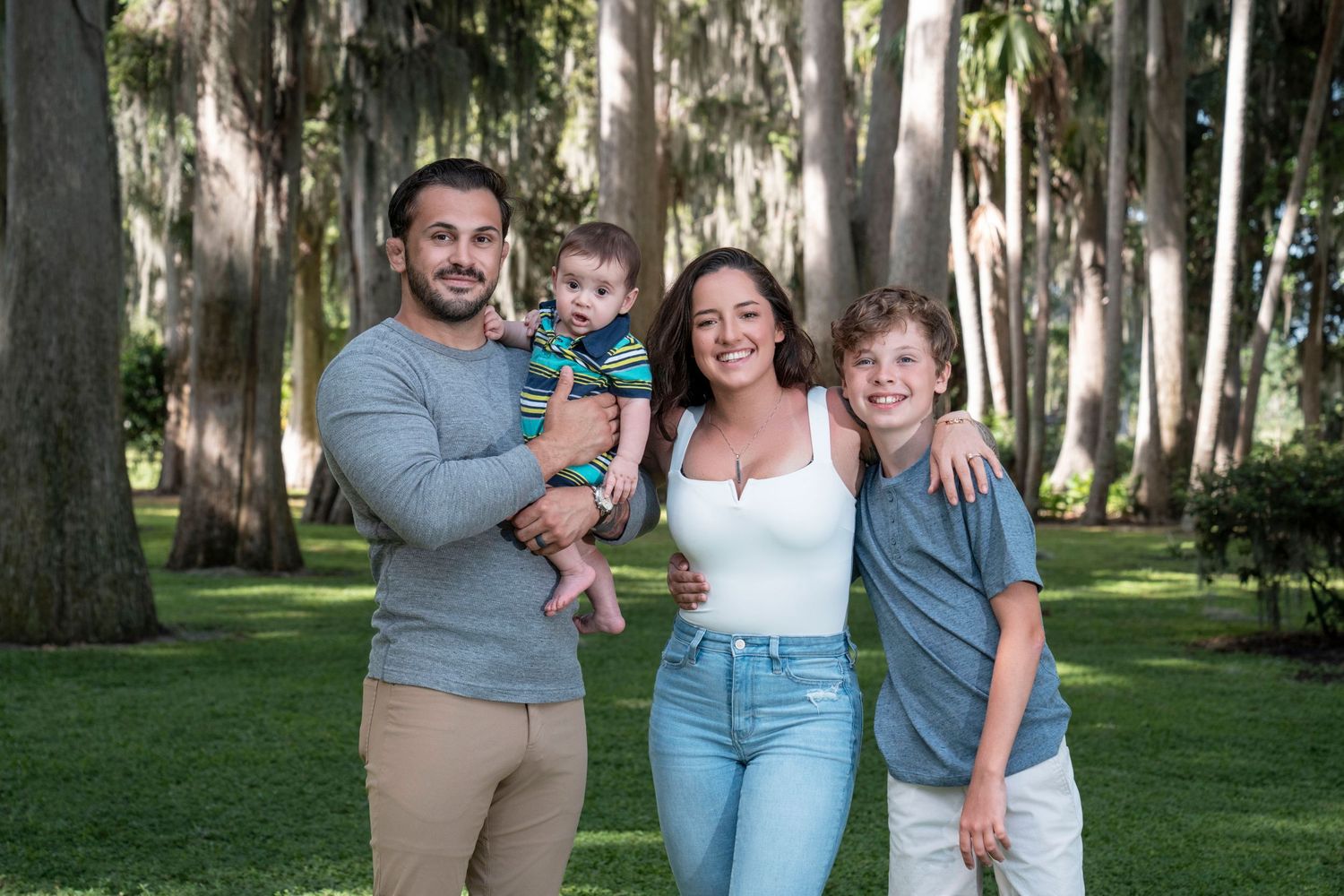 Family poses together in a park setting with tall trees and green grass in the background.