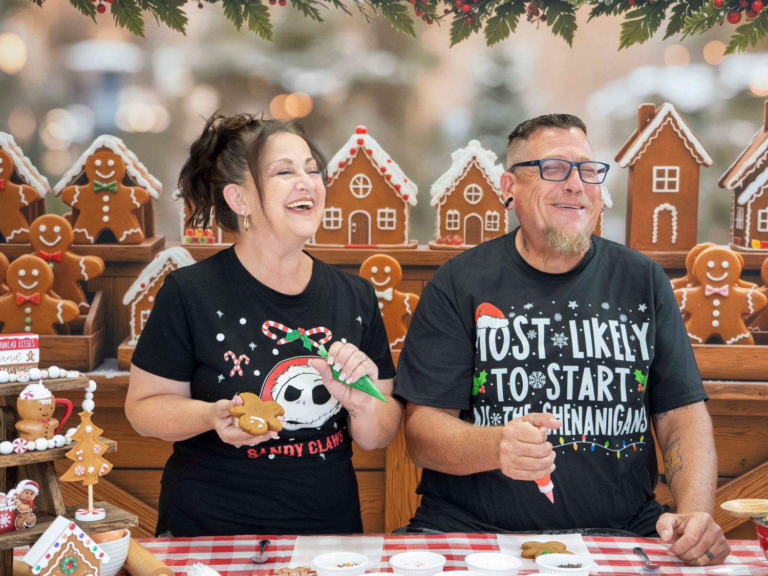 Two people in Christmas shirts laugh while decorating gingerbread cookies at a festive holiday table.