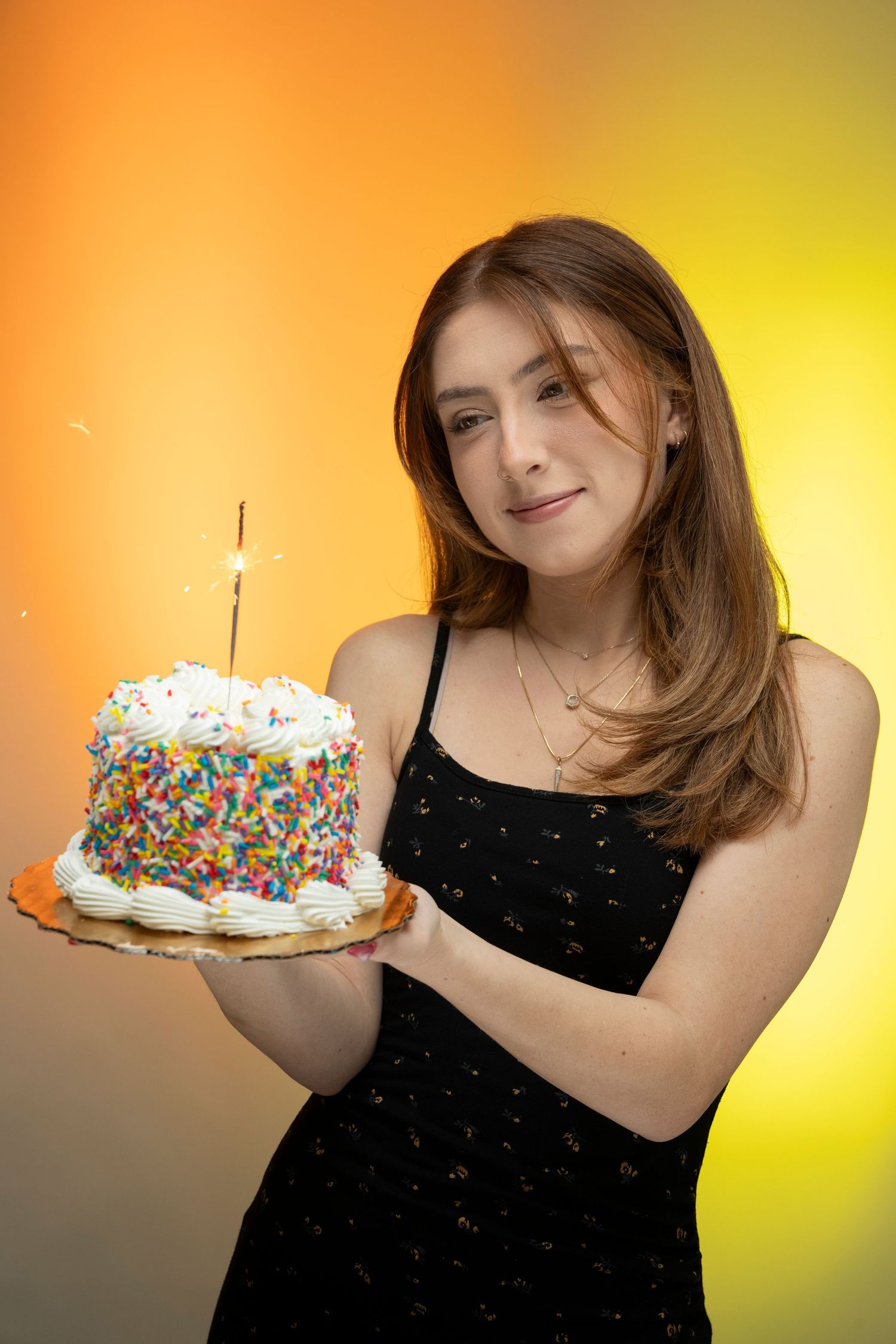 Young woman in black dress holding a colorful sprinkle birthday cake with a sparkler candle on orange background.