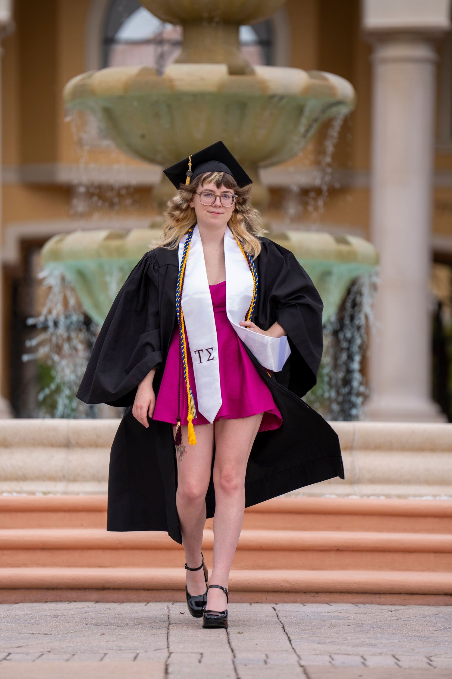 A smiling graduate in black cap and gown with white stole poses confidently in front of a fountain.