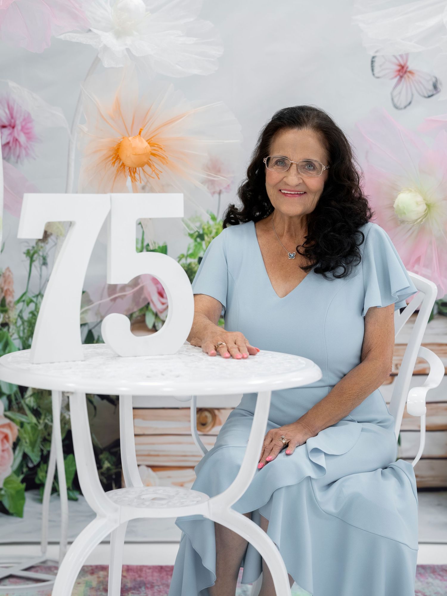 Elegant woman celebrating her 75th birthday seated at a white table with floral decorations and butterfly backdrop.