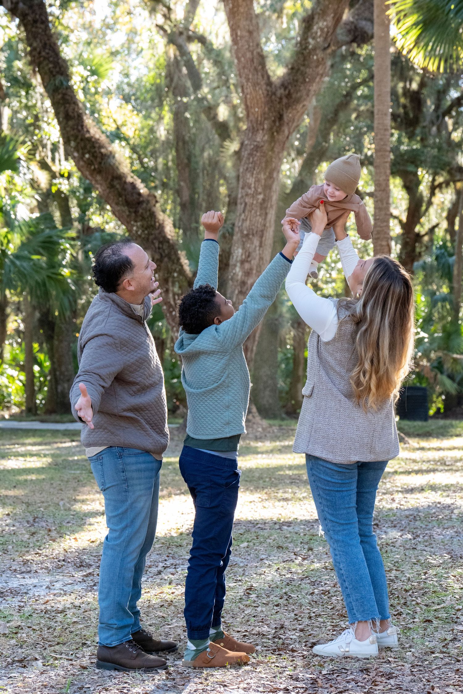 A joyful multigenerational family plays outdoors in a park, with a woman lifting a baby while others cheer.