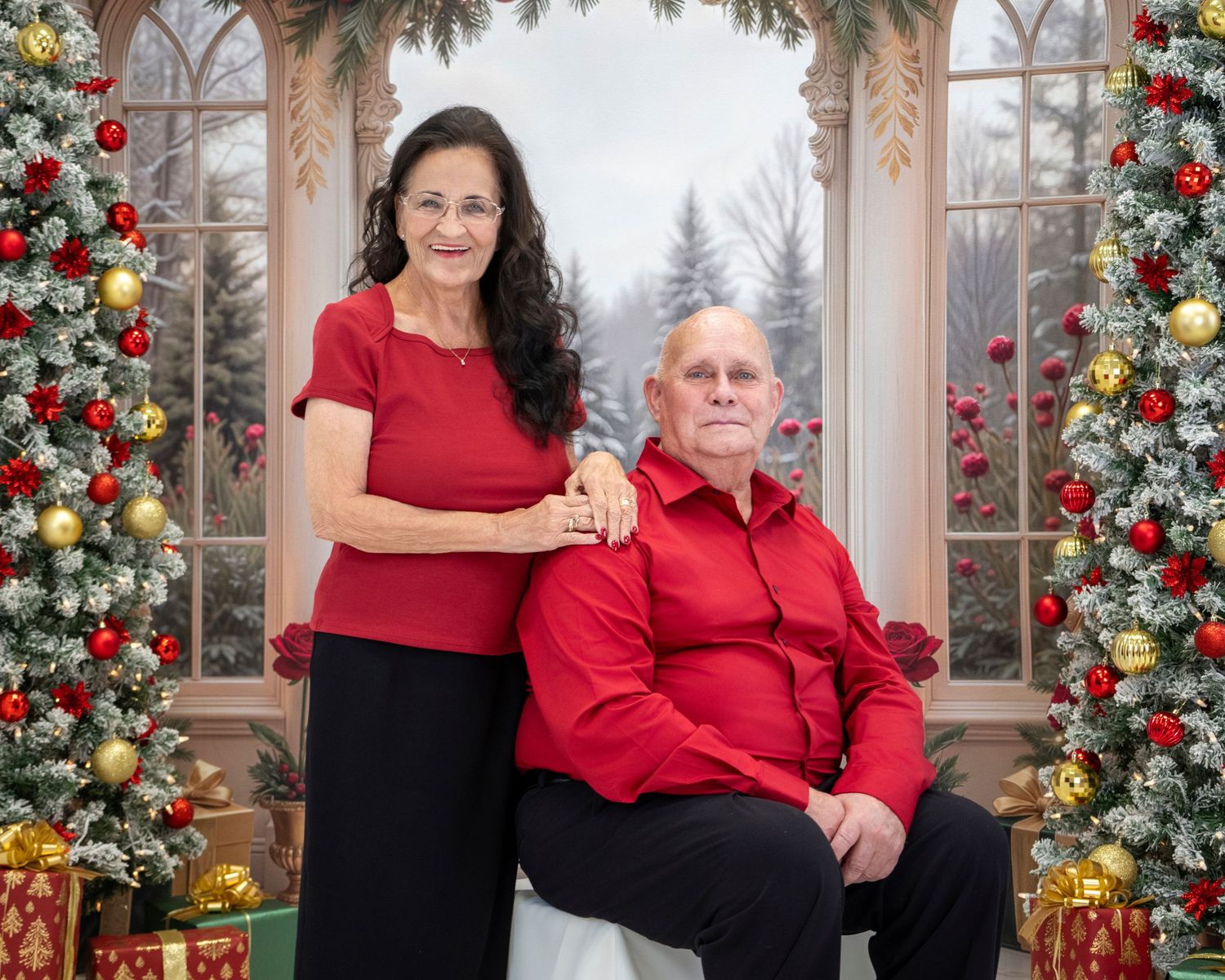 Couple in matching red outfits posing before a festive Christmas backdrop with decorated trees and gifts.