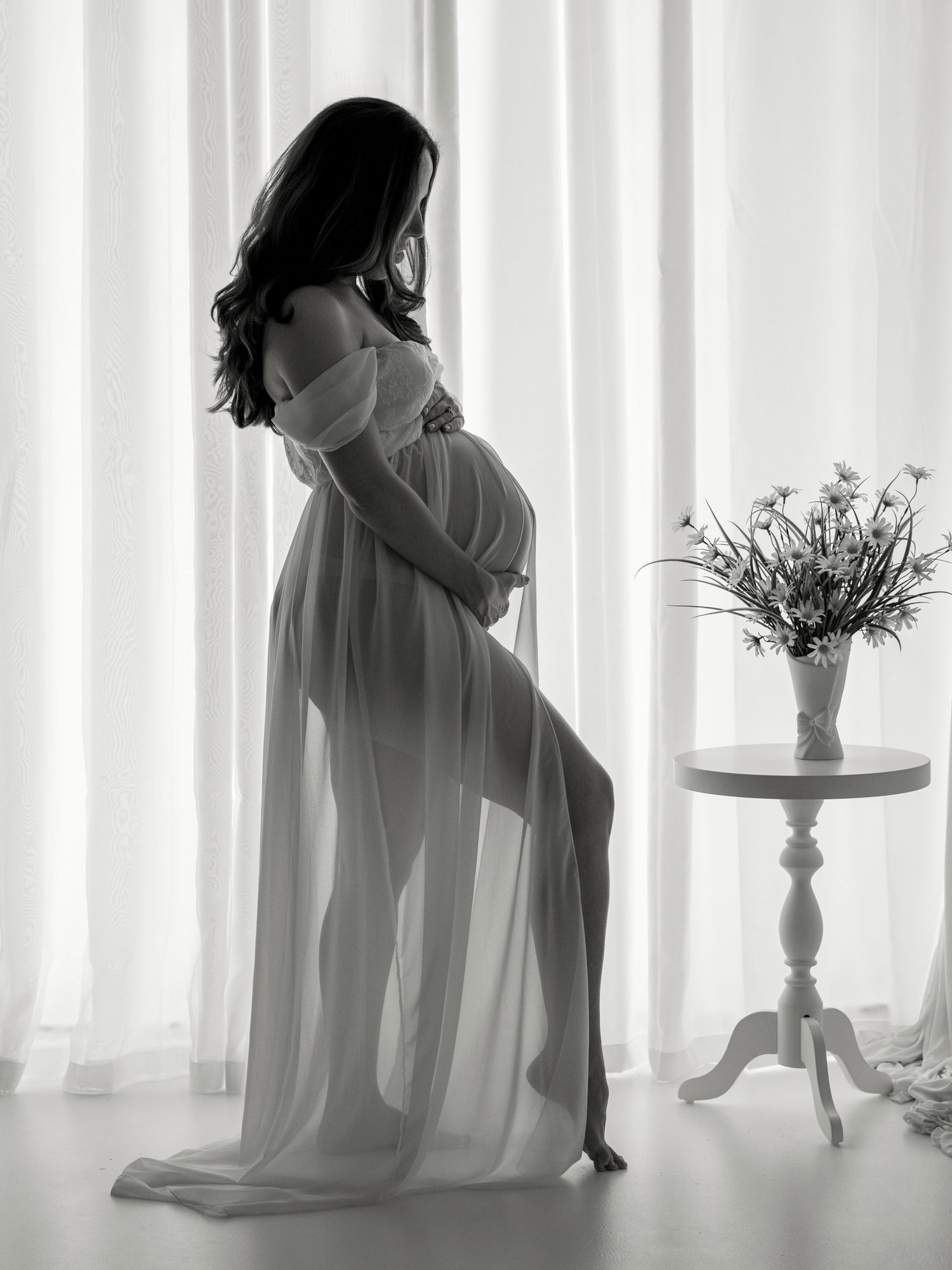 Black and white maternity photo of pregnant woman in sheer gown posing gracefully by curtains with floral arrangement nearby.