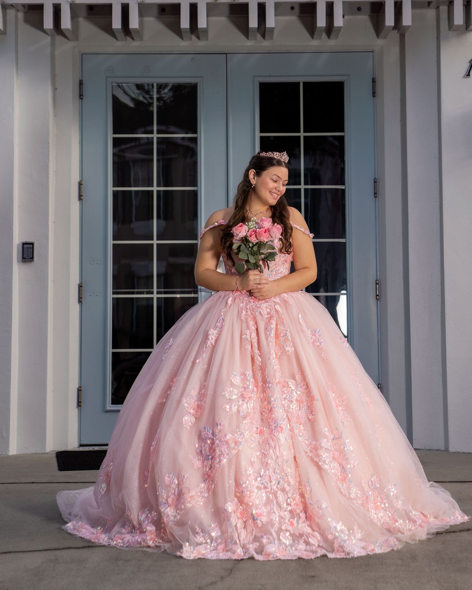 Young woman in pink floral ball gown holding bouquet, standing before blue French doors for quinceañera.