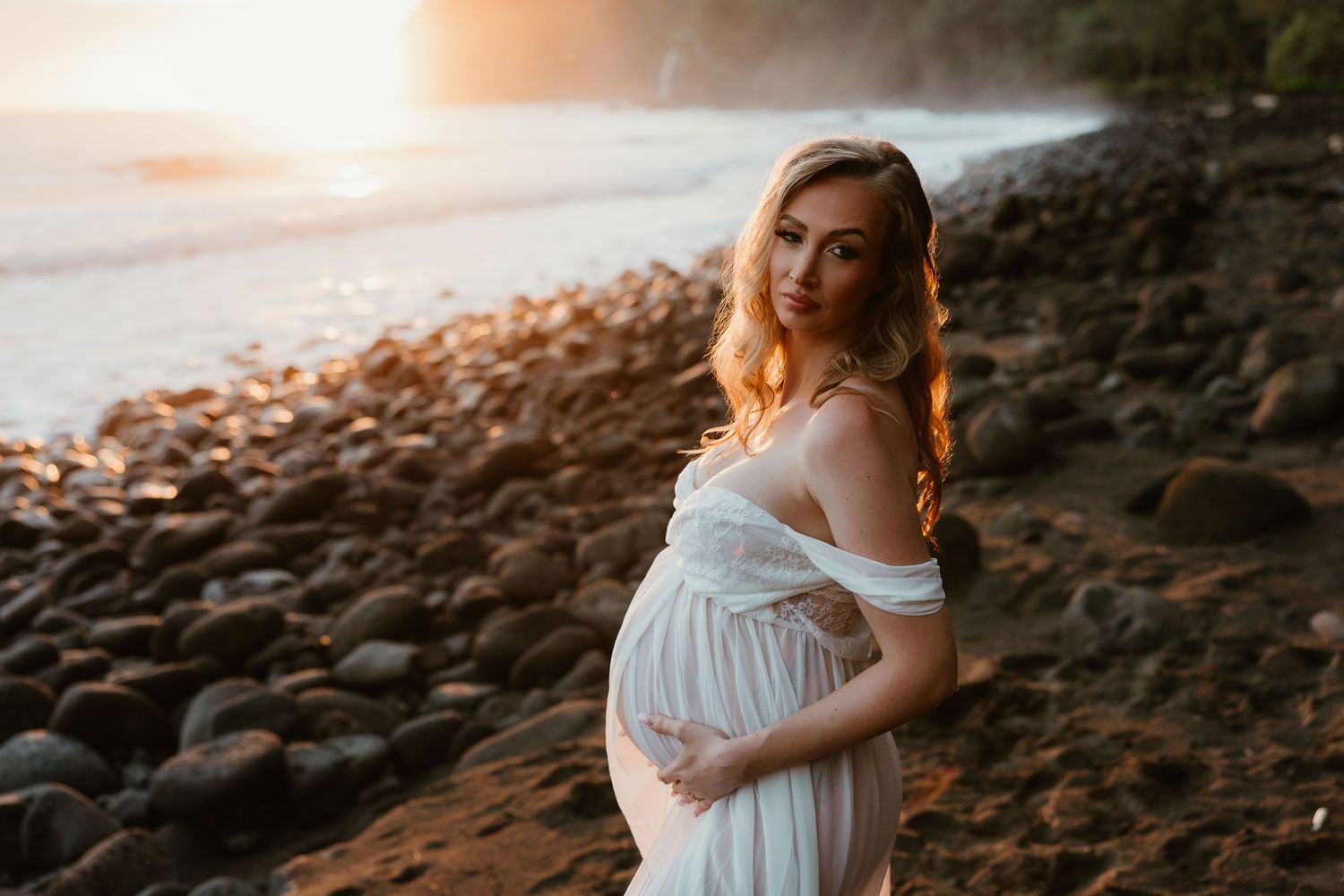 Pregnant woman in white dress holding baby bump on rocky beach at golden sunset.