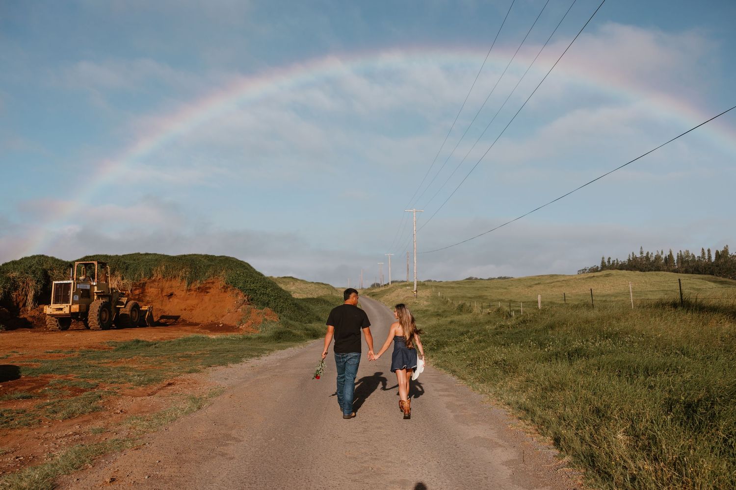 Couple walking hand-in-hand down a rural road under a vibrant rainbow with green hills and blue sky.